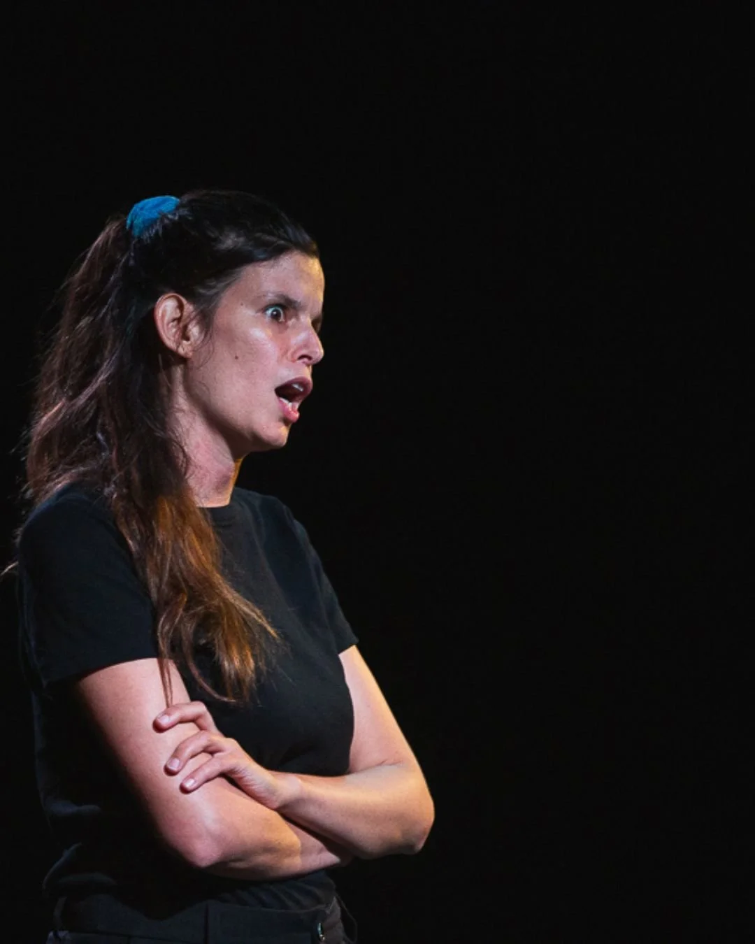 Une femme avec des cheveux longs, portant un t-shirt noir, a l'air surprise ou en colère, sur un fond noir.