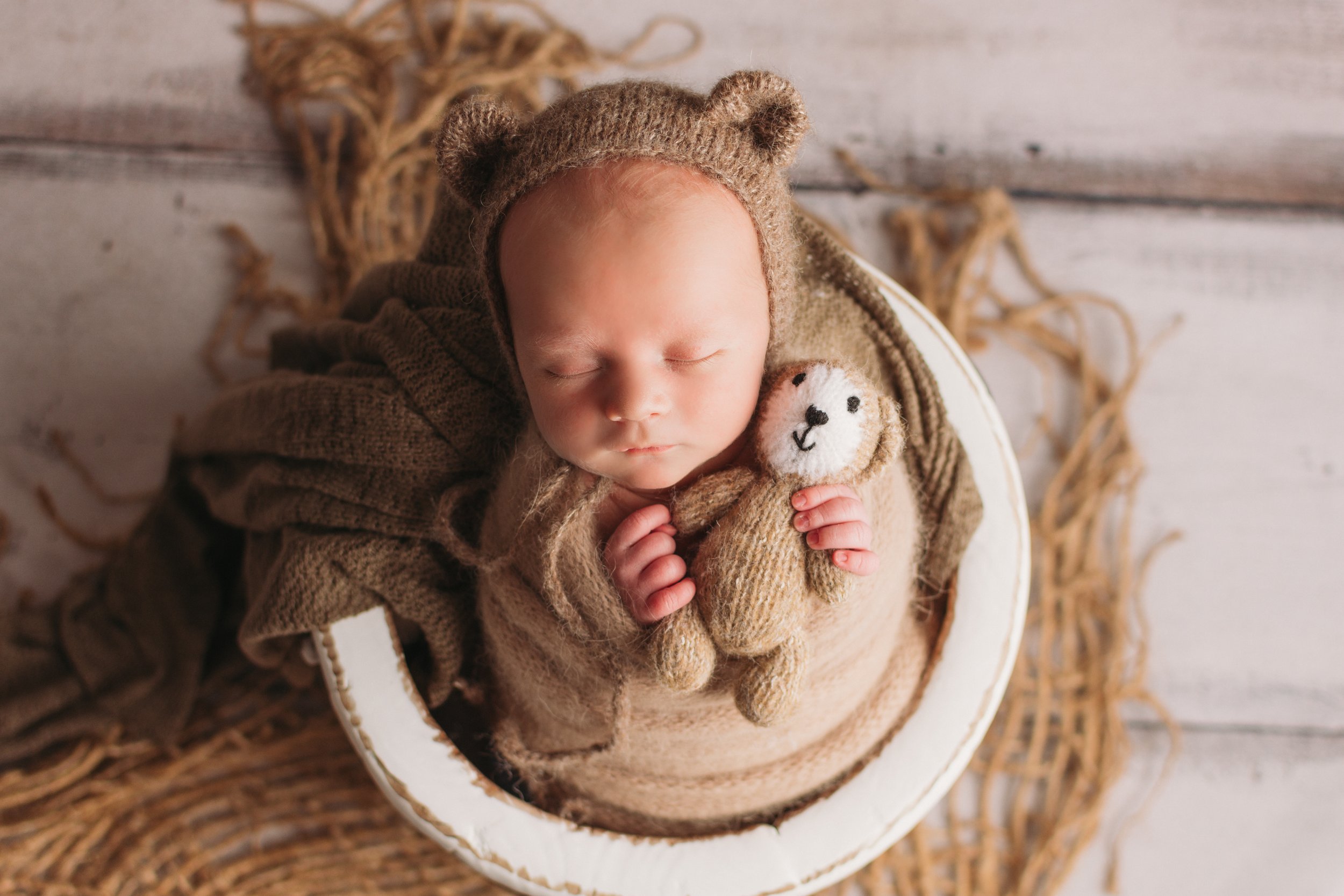 newborn in bucket holding teddy bear wearing bonnet