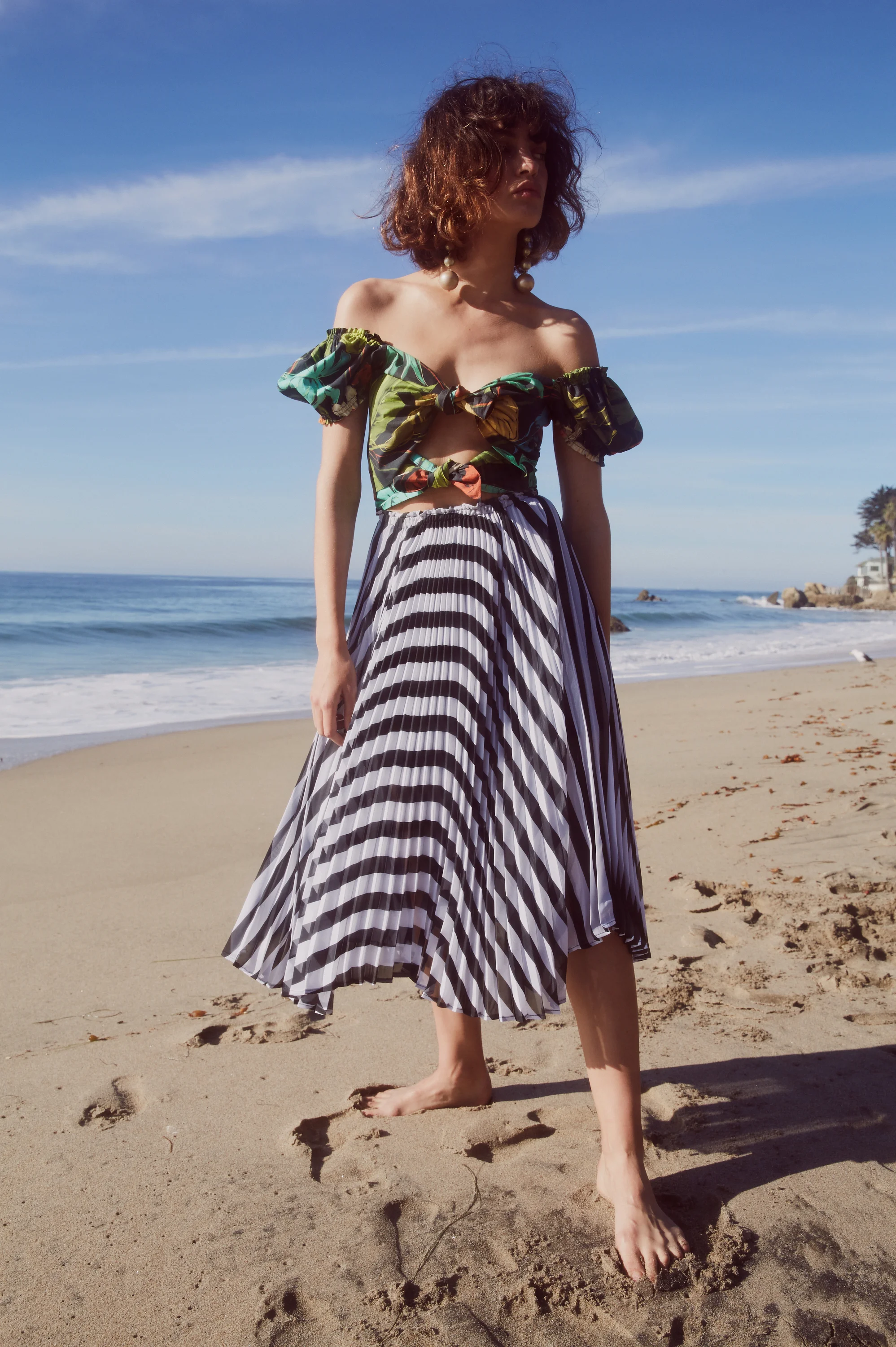 Model stands on a beach in a mixed print off the shoulder dress with black floral print bodice and horizontal black and white stripe skirt