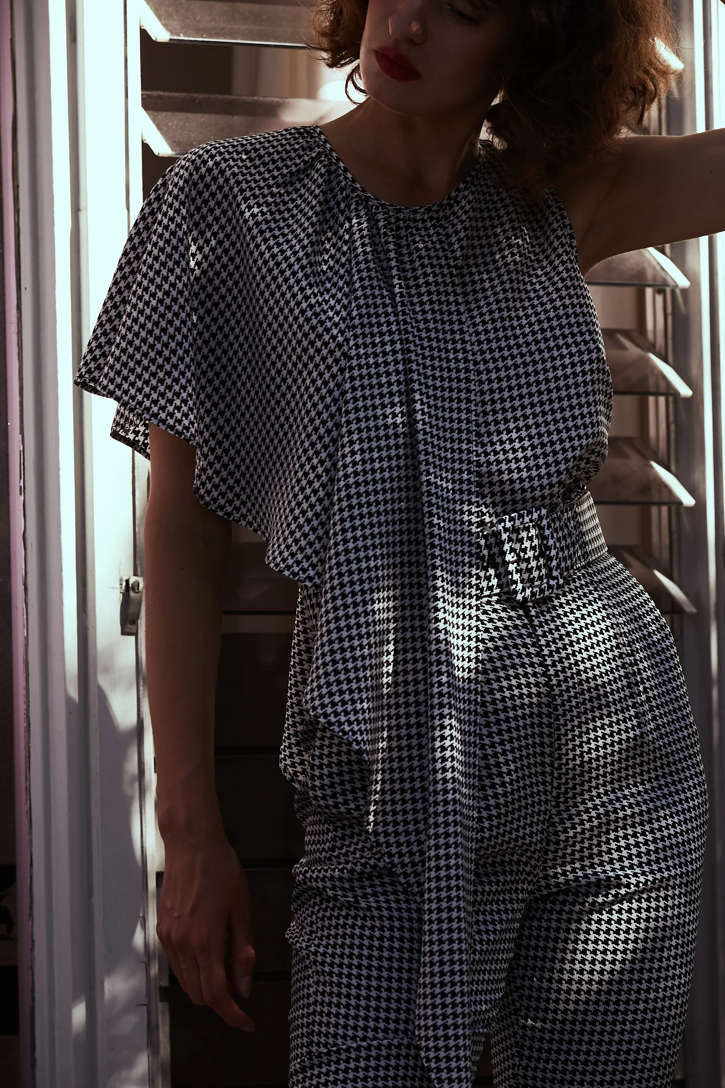 Model stands in sleeveless blouse in mini houndstooth print with asymmetric front ruffle overlay