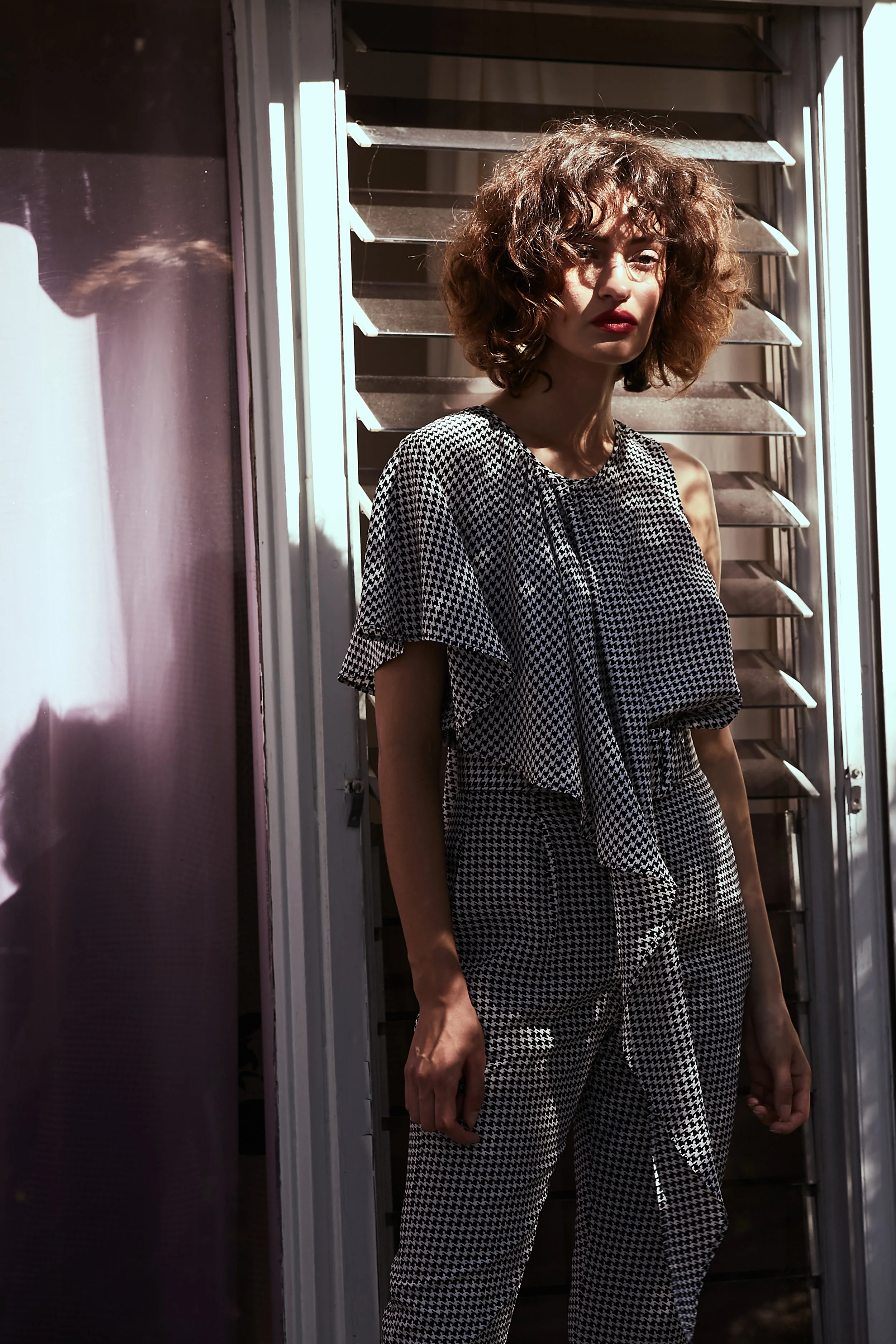 Model stands in shadows in sleeveless blouse in mini houndstooth print with asymmetric front ruffle overlay