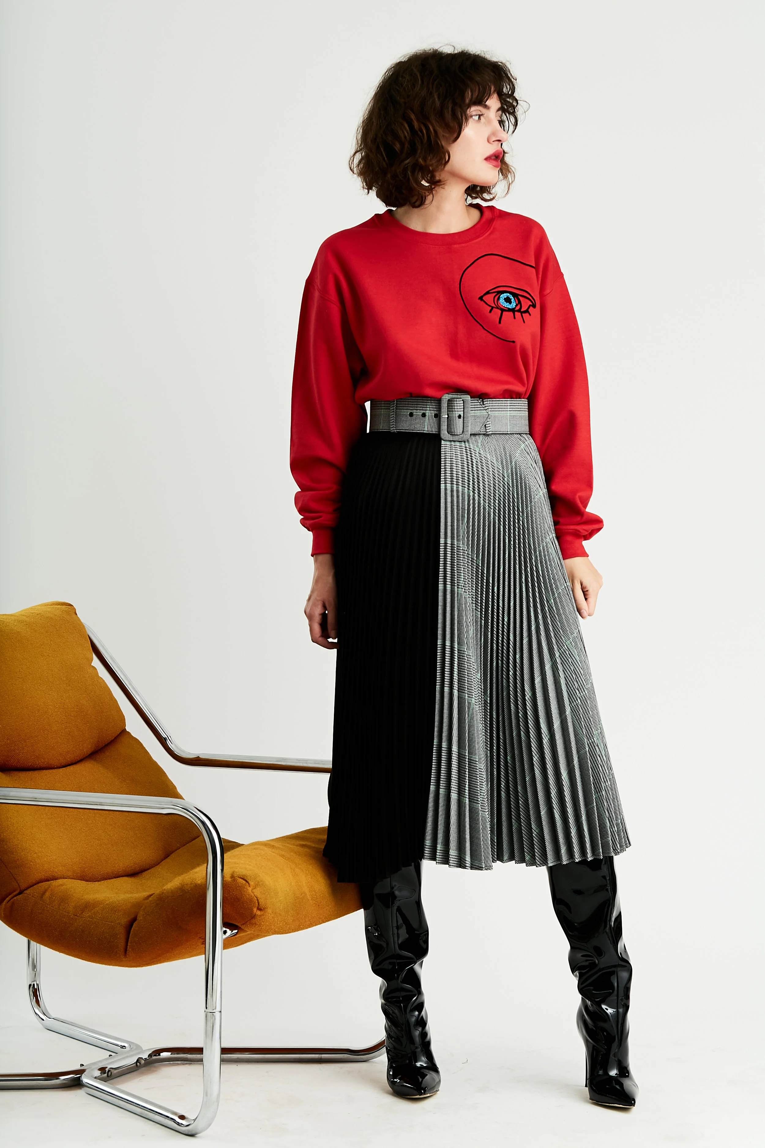 Model stands in red crewneck sweatshirt with blue eye flocked graphic and color block pleated skirt half plaid half black