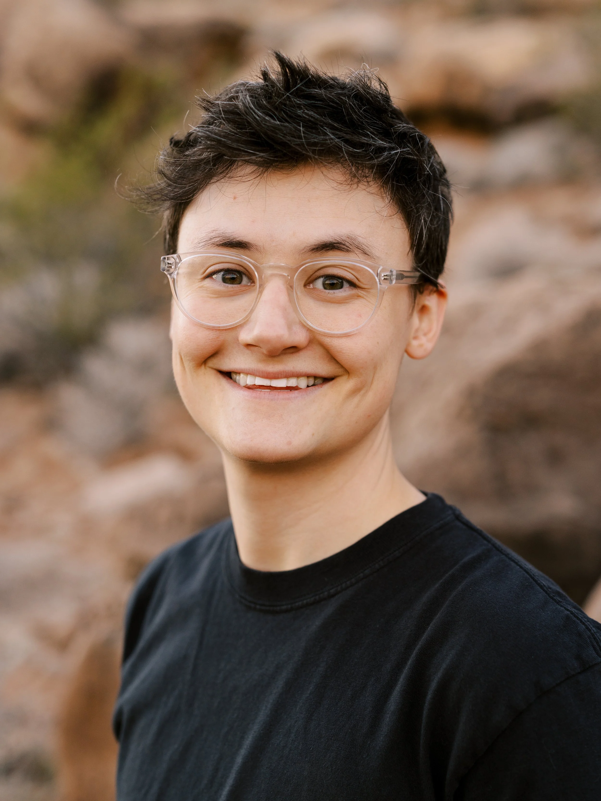 Headshot of Jen with short dark hair and clear-framed glasses, smiling, outdoors