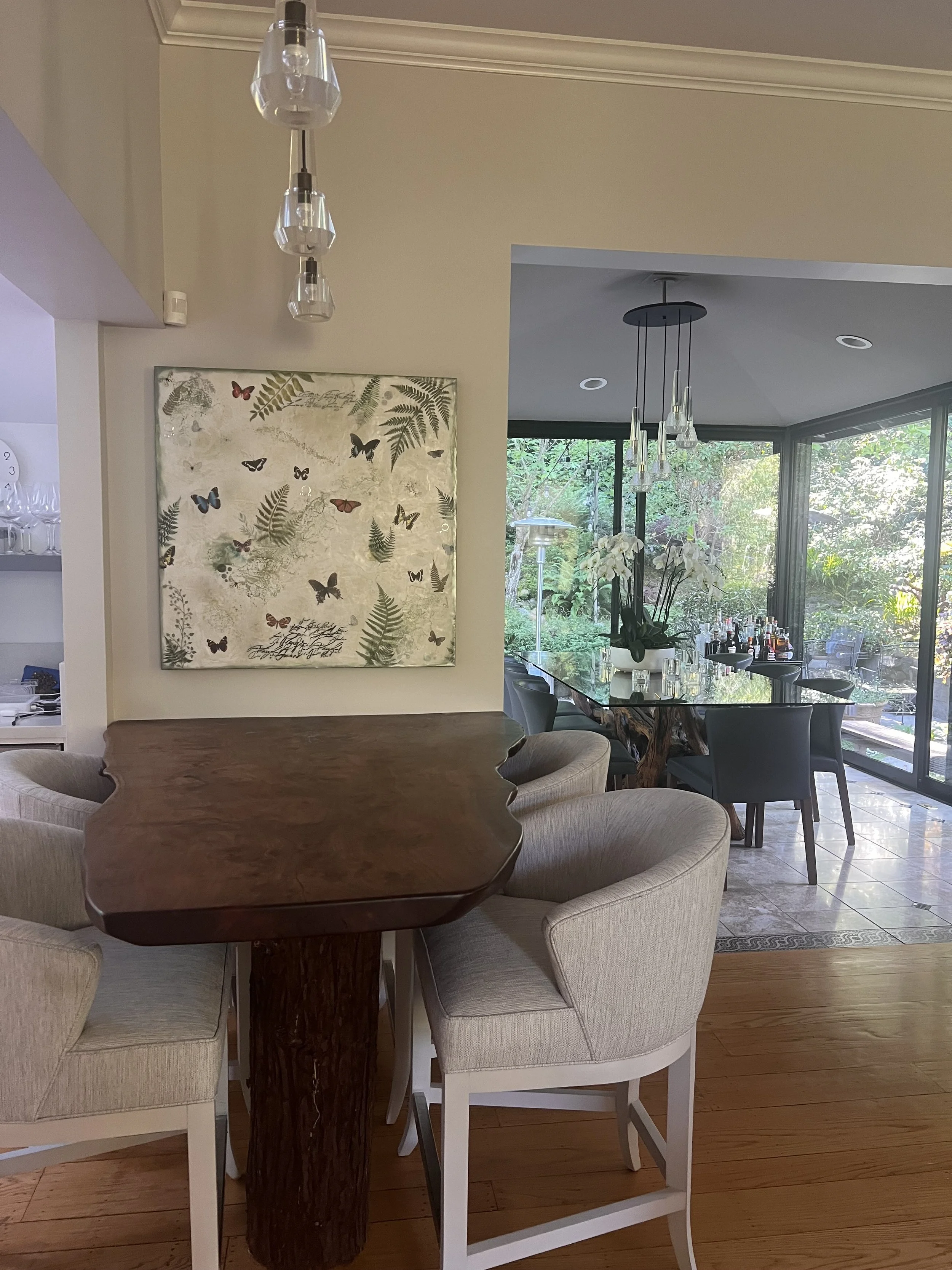 Interior view of a dining room with a wood table, upholstered chairs, and a view of a garden through large windows, decorated with plants and art on the wall.