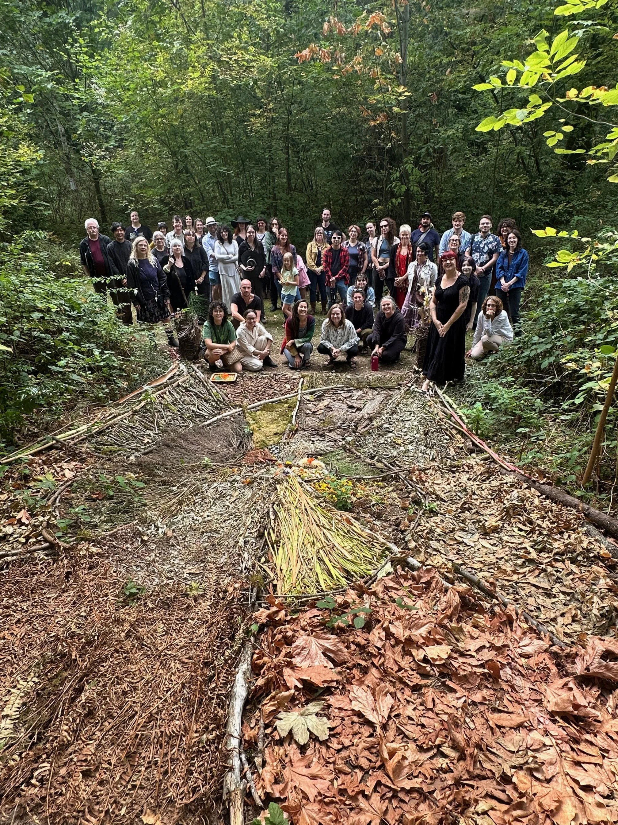 A large group of people gathered in a forested area with fallen leaves, standing on a trail, with some sitting or kneeling at the front. There are trees and greenery surrounding them.