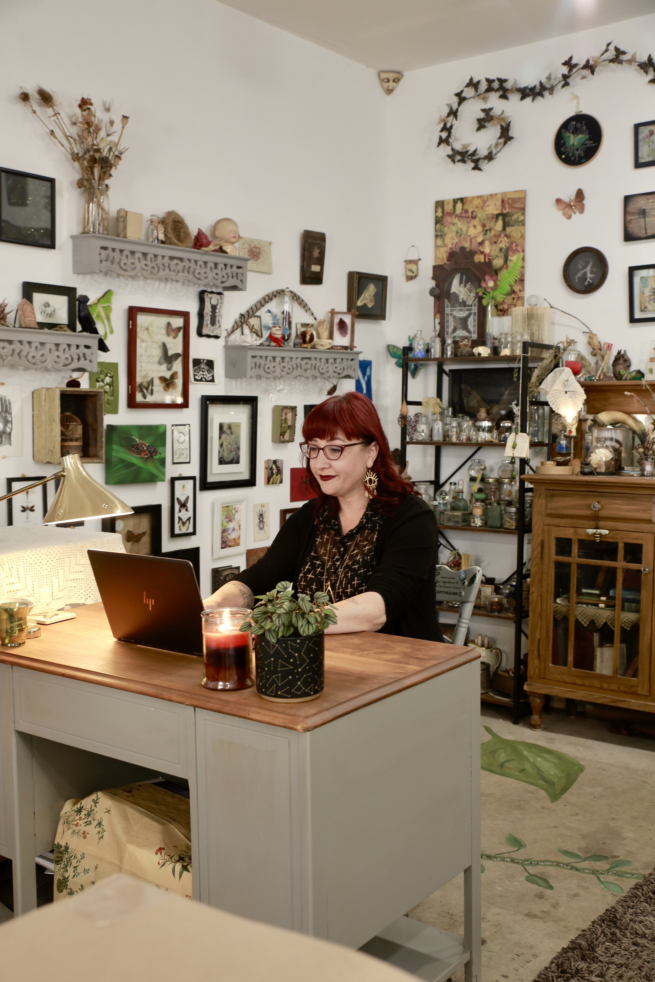A woman with red hair, glasses, and earrings is sitting at a desk working on a laptop in a room decorated with numerous picture frames, artworks, and decorative objects.