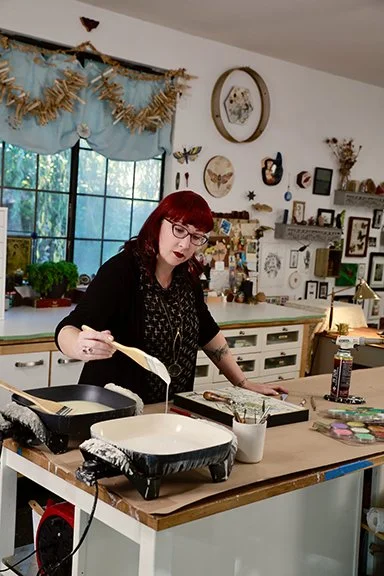 Person with red hair and glasses cooking with a spatula in a home kitchen decorated with framed pictures, butterflies, and a window with blue curtains.