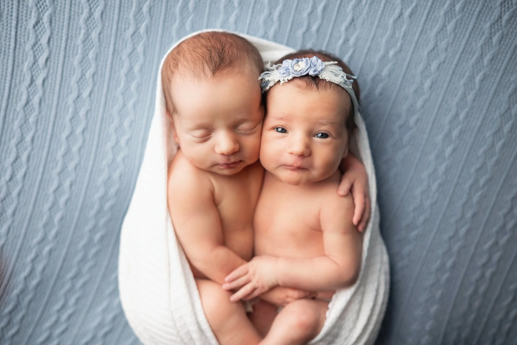 Two newborn babies wrapped in a white blanket, lying on a soft gray surface, with one baby wearing a floral headband.
