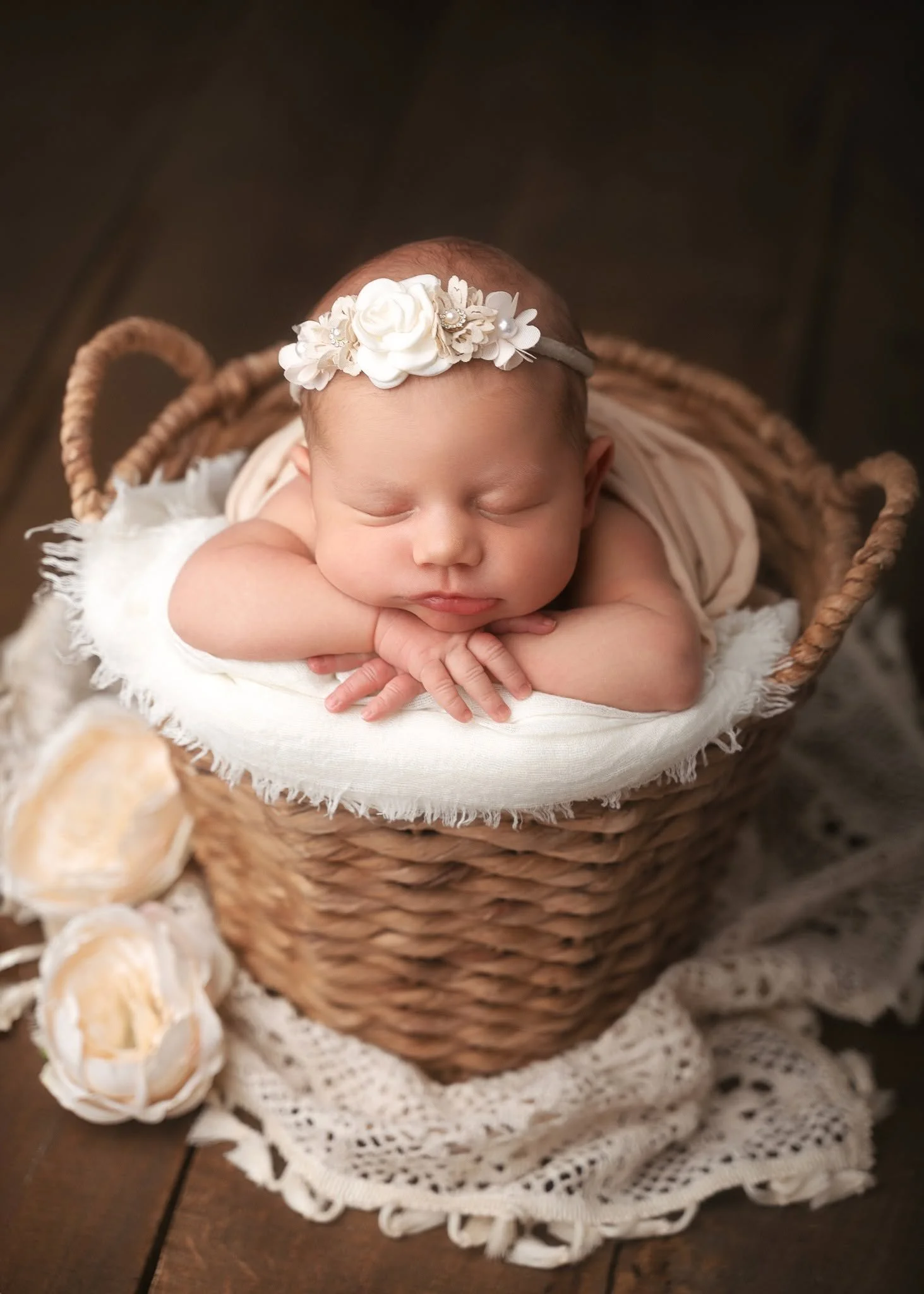 A sleeping newborn baby with a floral headband in a wicker basket, wrapped in a soft blanket with lace trim, beside some roses.