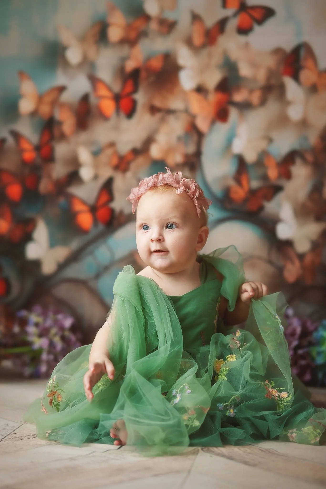 A young girl wearing a green dress with a pink flower crown is sitting on the floor in front of a butterfly mural backdrop.