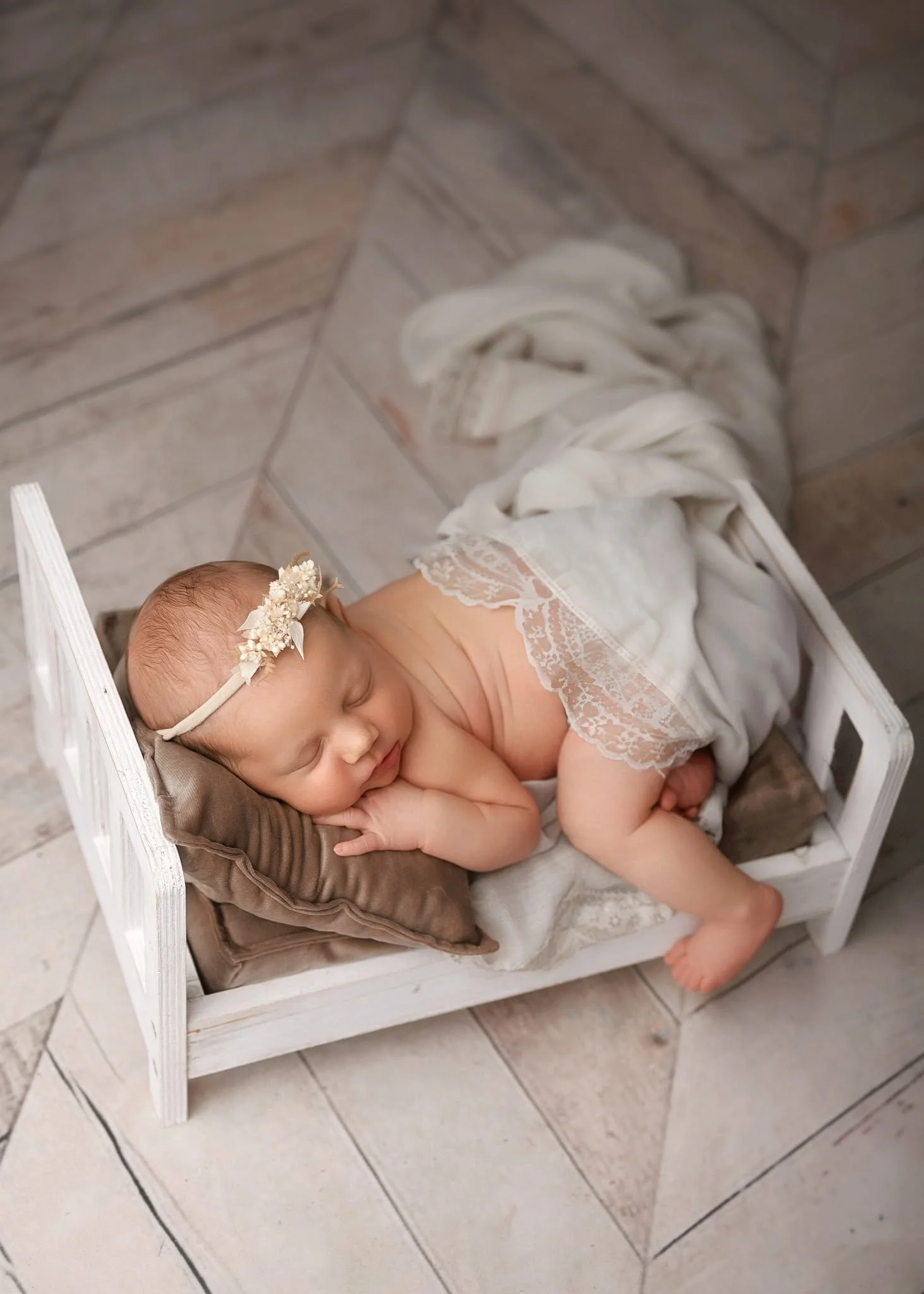 A sleeping baby girl lying on a small white wooden bed, wearing a lace outfit and a flower headband, on a tiled floor.