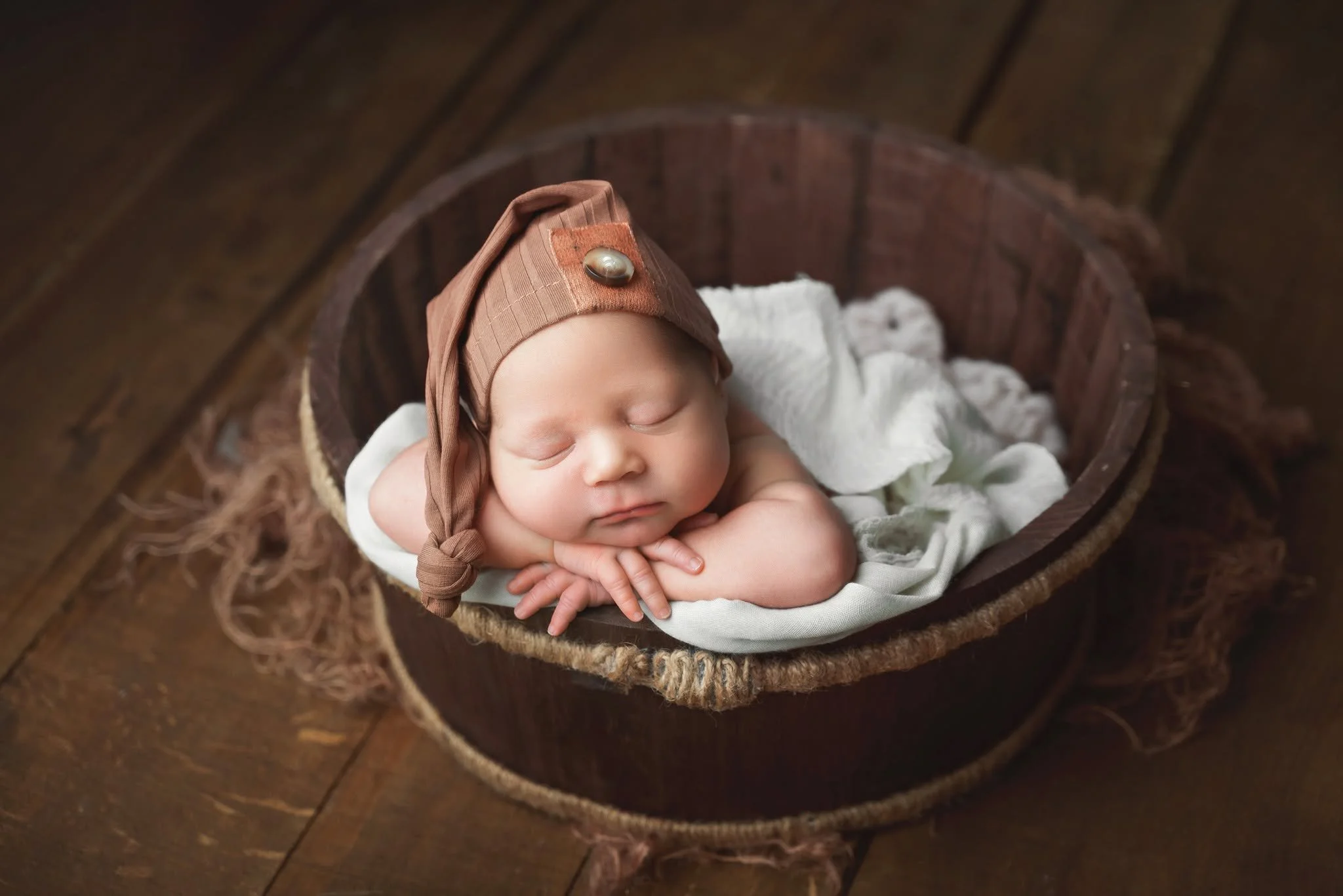 A sleeping baby in a brown hat with a pearl button, lying on a white blanket inside a rustic wooden cradle.