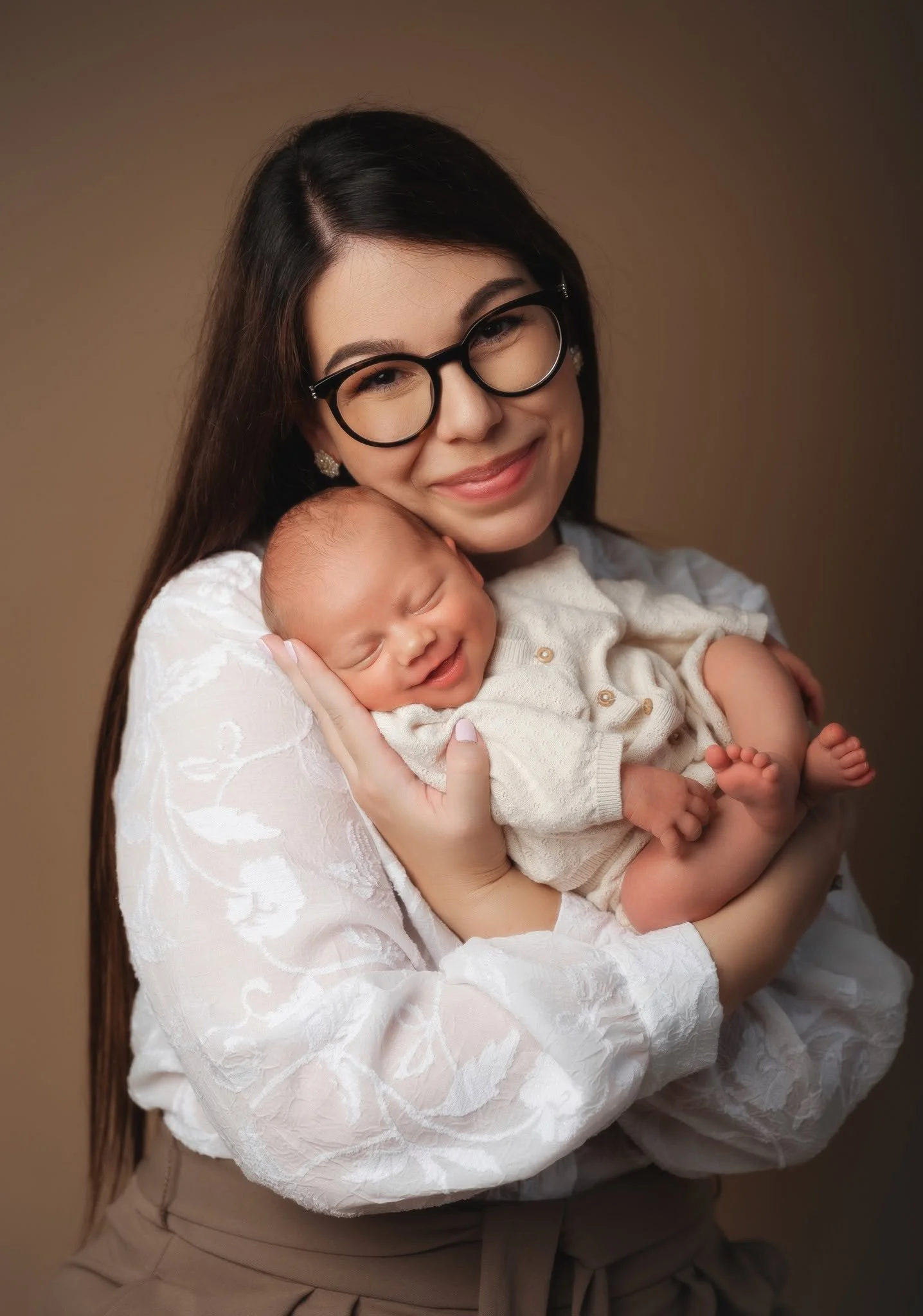 A woman wearing glasses and earrings holding a sleeping baby dressed in a light-colored outfit with buttons, against a brown background.