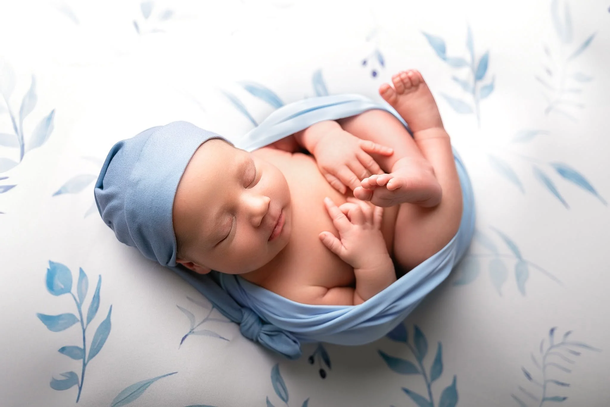 A sleeping newborn baby wearing a blue bonnet, lying on a white sheet with blue leaf patterns, wrapped in a blue swaddle.