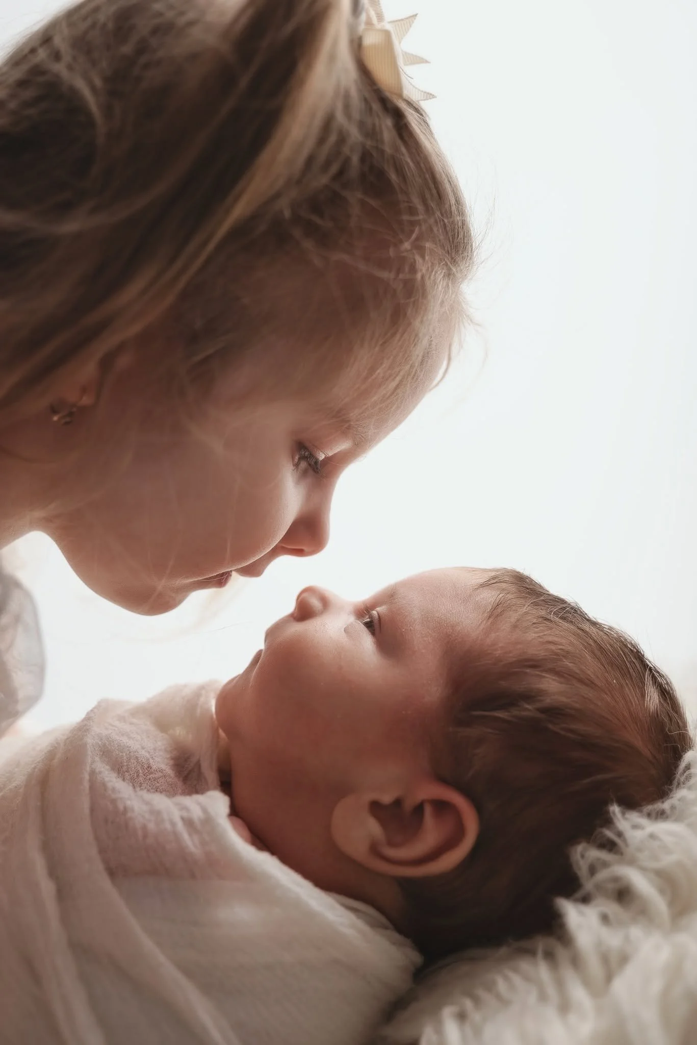 A woman and a baby are close together, with their faces nearly touching. The woman has light brown hair with a bow, and the baby has light brown hair and is lying on a soft, white surface.