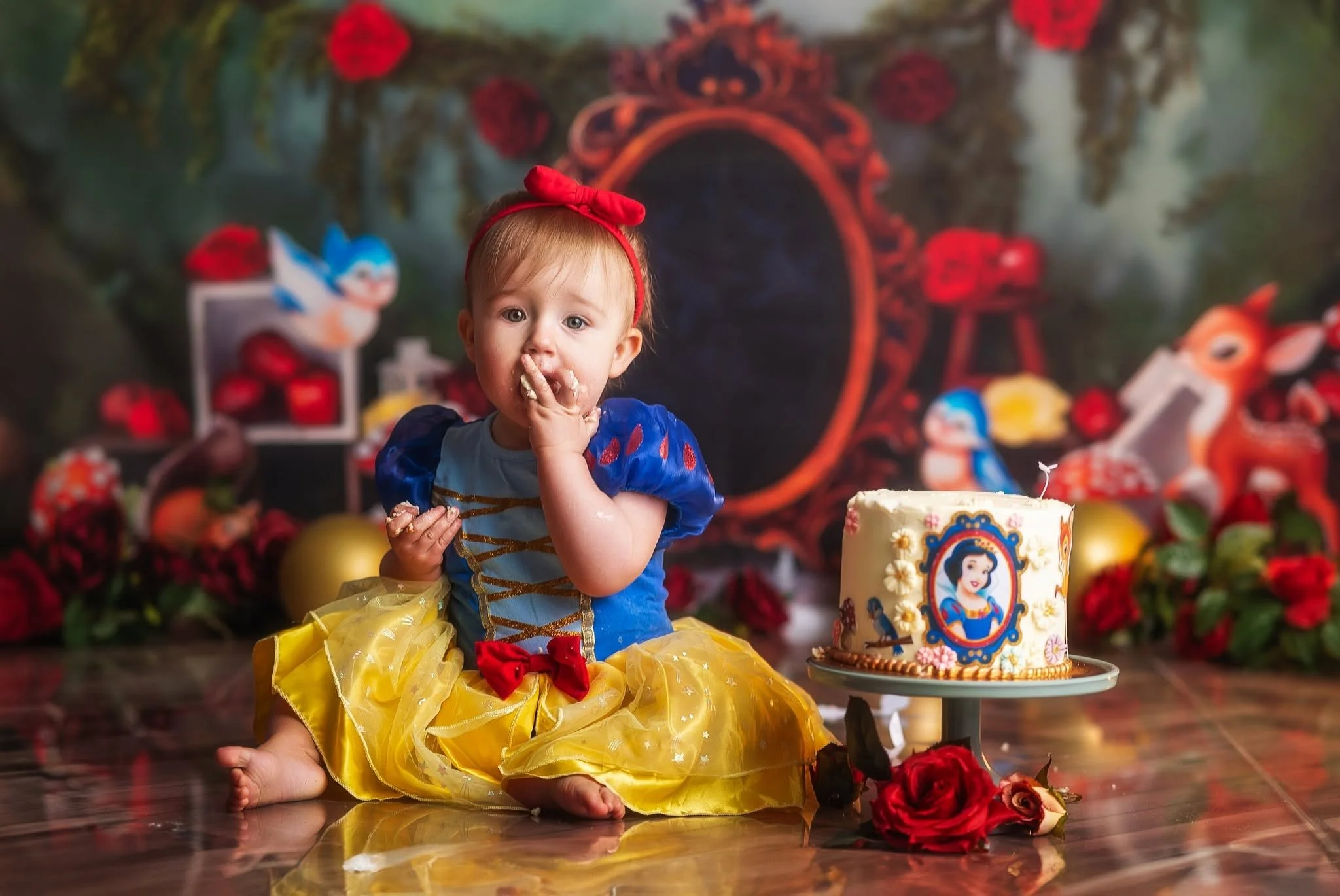 A young girl dressed as Snow White sitting on the floor with her hand on her face. She has a birthday cake decorated with Snow White's image in front of her, with red roses and balloons in the background.