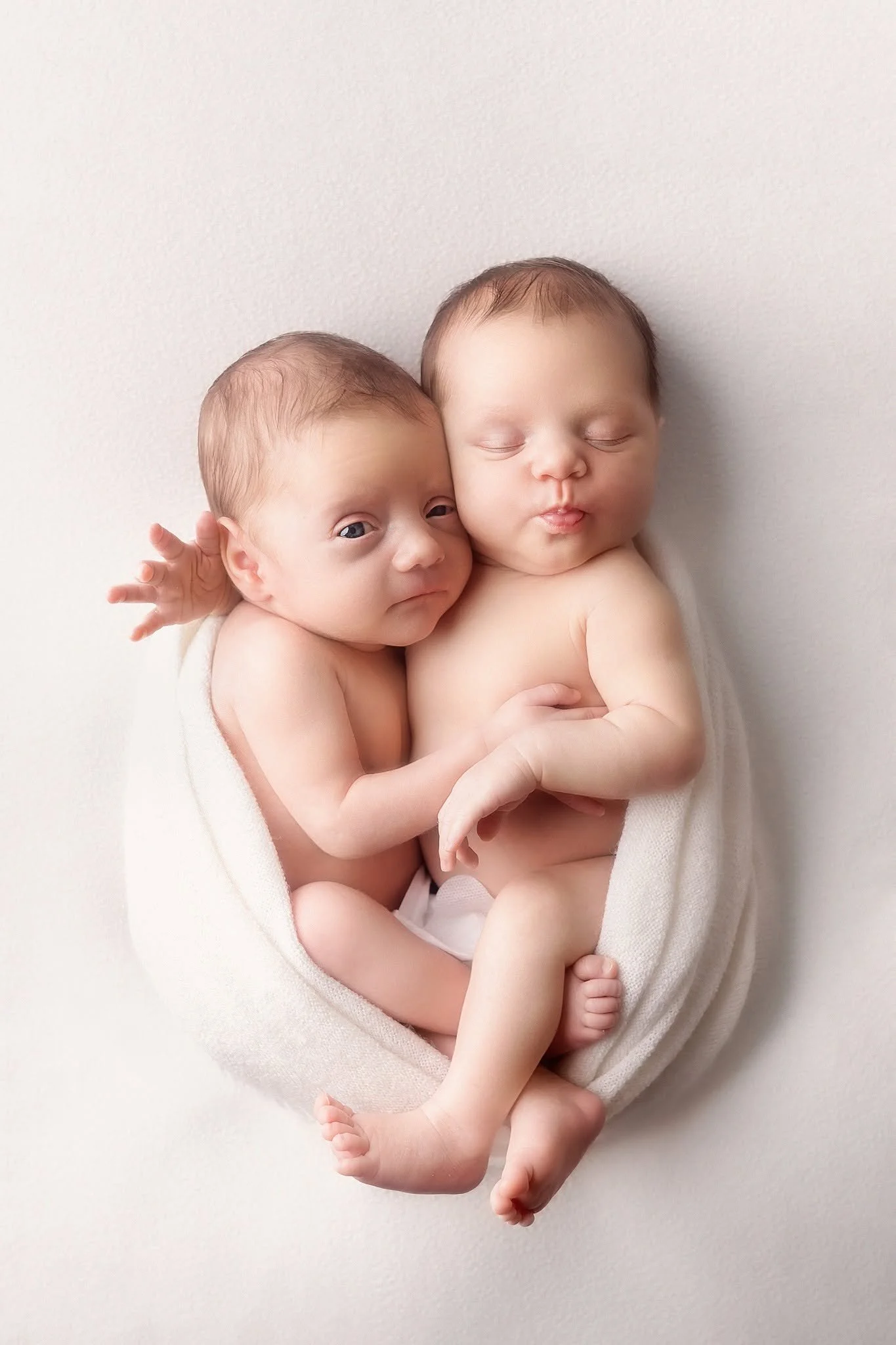 Two newborn babies, one awake with eyes open and the other with eyes closed, embracing each other wrapped in a white cloth against a light background.
