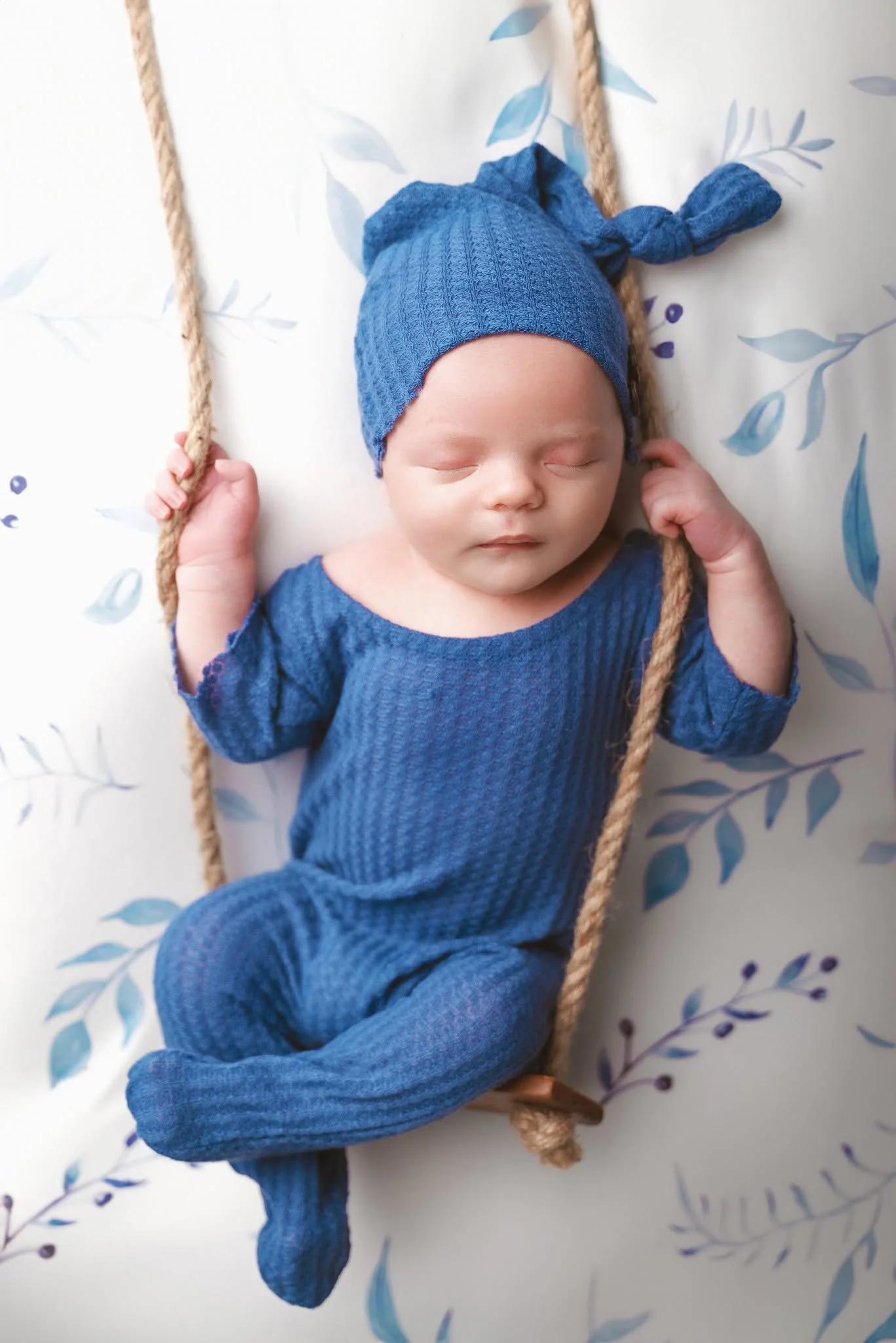A baby sleeping while lying on a white sheet with a leafy pattern, wearing a blue knit hat with a knot and matching blue outfit, holding a thick rope with both hands.