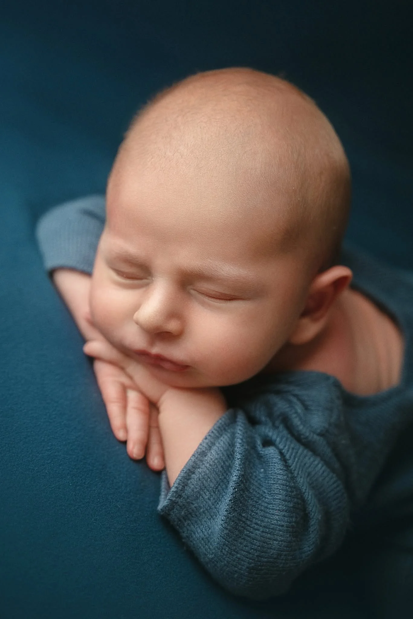 A young baby with a shaved head, sleeping peacefully with hands folded under their chin, dressed in a gray long-sleeve shirt, lying on a dark blue surface.