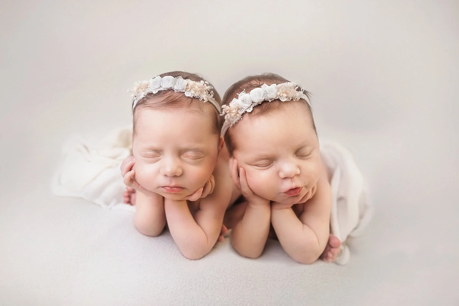 Two sleeping babies with headbands, lying on their stomachs with their hands under their chins on a white surface.