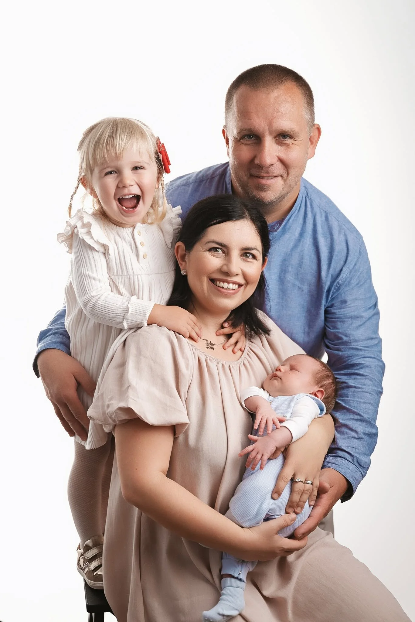 A multi-generational family portrait of a man, woman, two young girls, and a baby, all smiling and closely gathered against a plain white background.