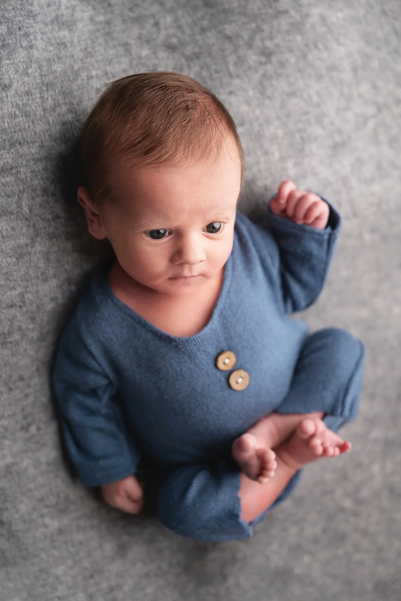 A baby with blue eyes and light brown hair lying on a gray surface, wearing a dark blue cardigan with three wooden buttons.