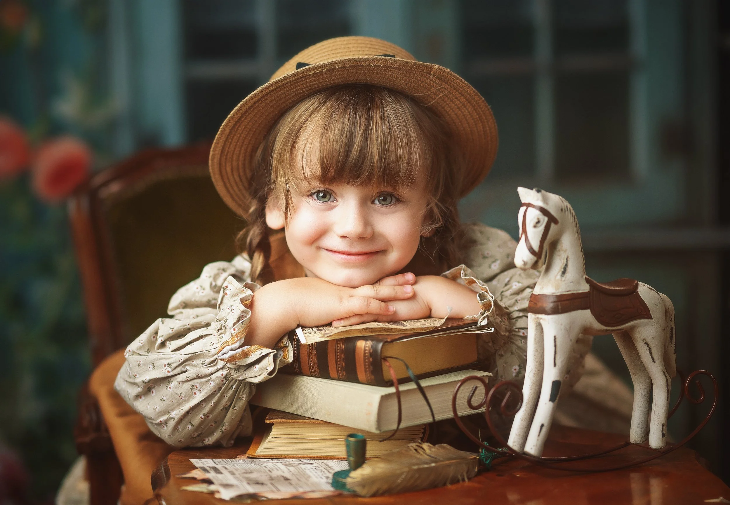 A young girl with curly hair and blue eyes, wearing a straw hat, resting her chin on folded hands over a stack of books, sitting on a wooden chair in a cozy indoor setting, with a vintage rocking horse in front of her.
