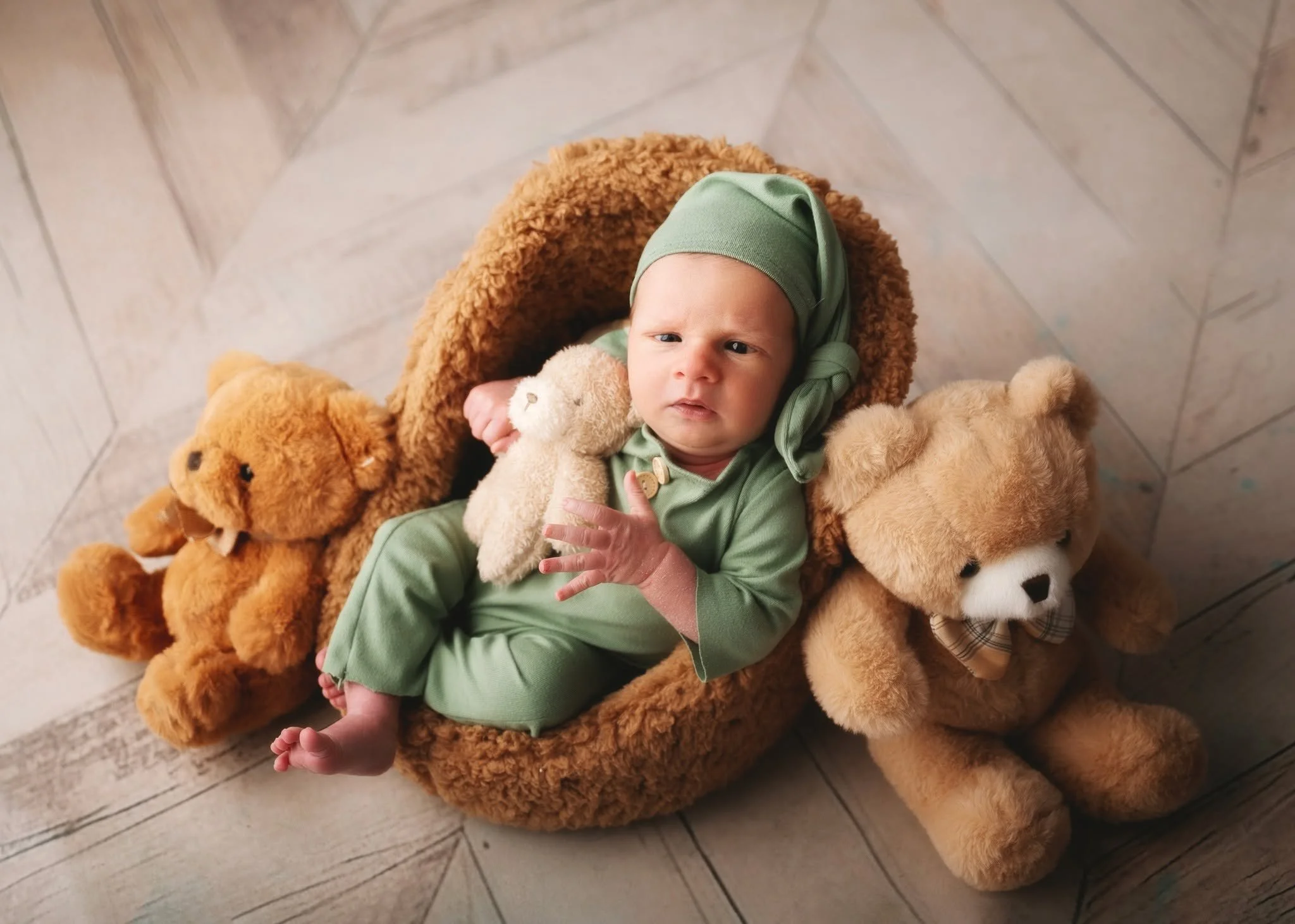 A baby is lying on a large teddy bear, wearing a green hat and matching outfit, surrounded by teddy bears on a wooden floor.