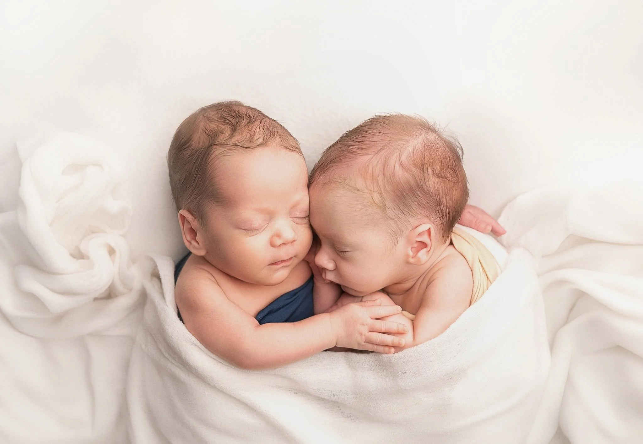 Two newborn babies sleeping closely together on a white blanket.