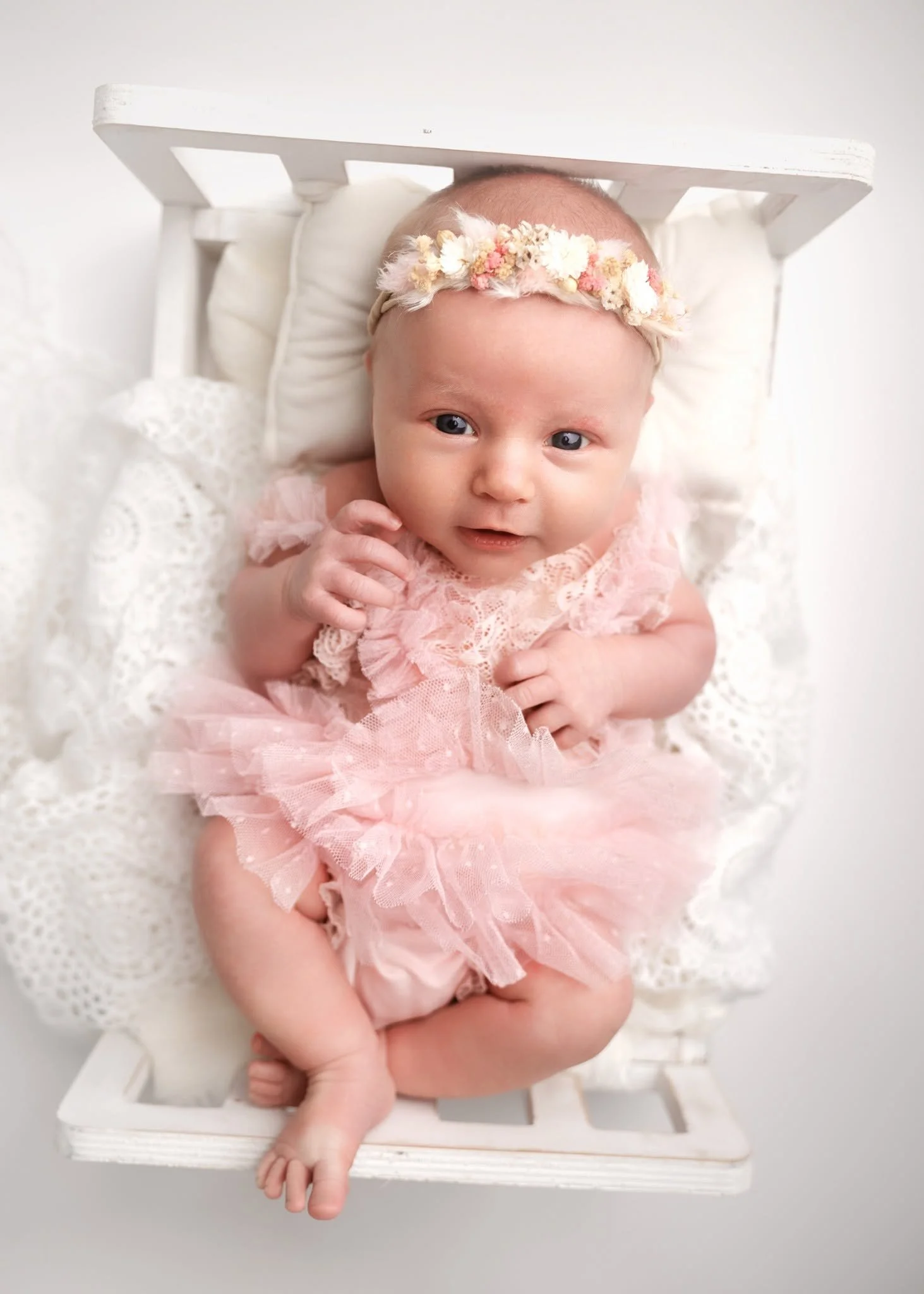 A baby girl with blue eyes wearing a pink lace dress and a floral headband, lying in a white cradle with lace and wood elements.