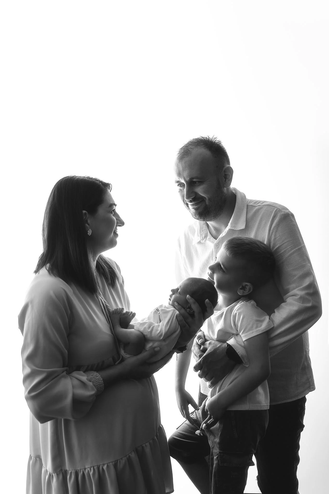 A family of five, including two adults, a teenage girl, a young boy, and a newborn, are engaging in a tender moment indoors with a bright background.