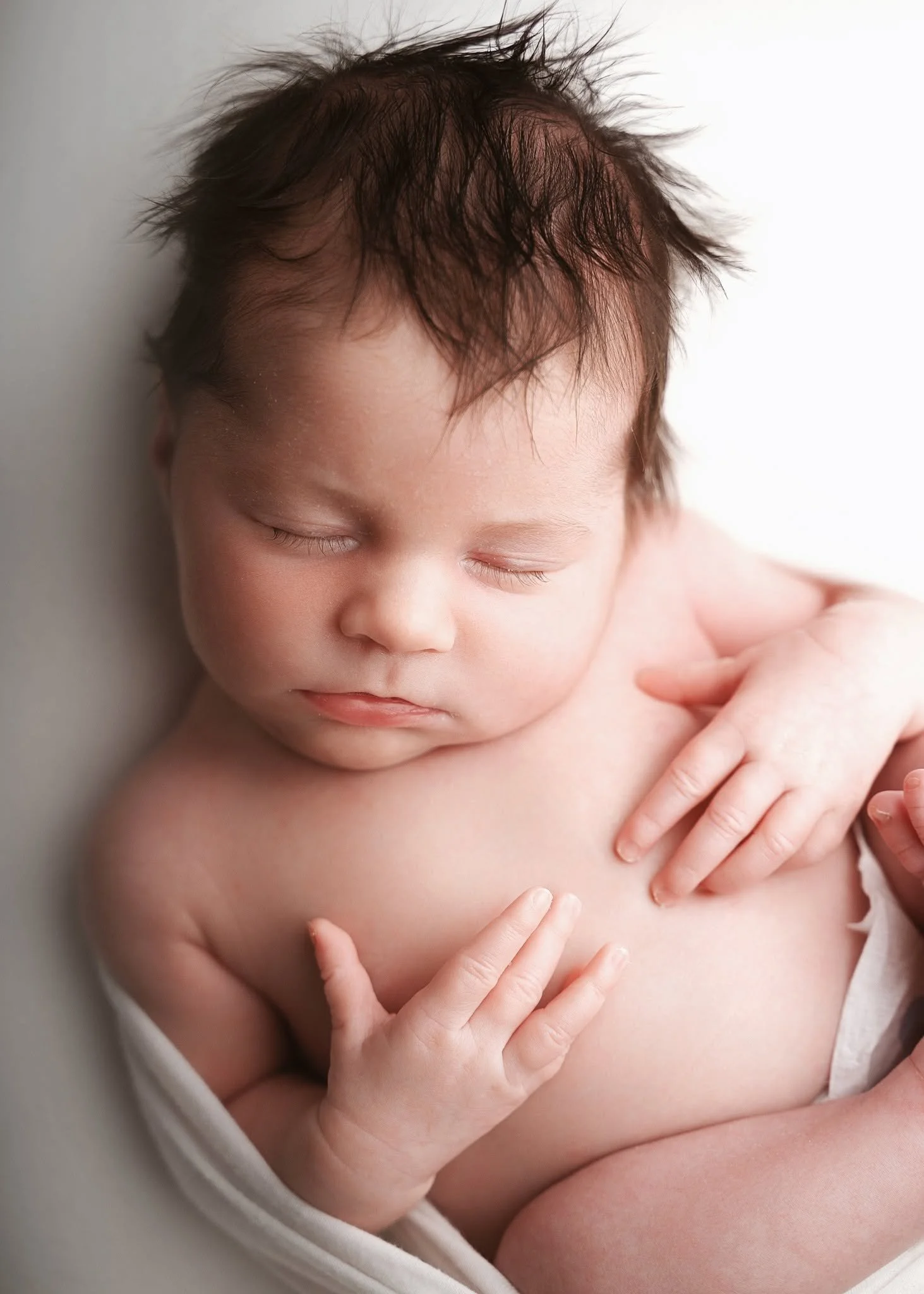 Close-up of a sleeping baby with tousled hair, resting head against a white background, with gentle light illuminating the face.