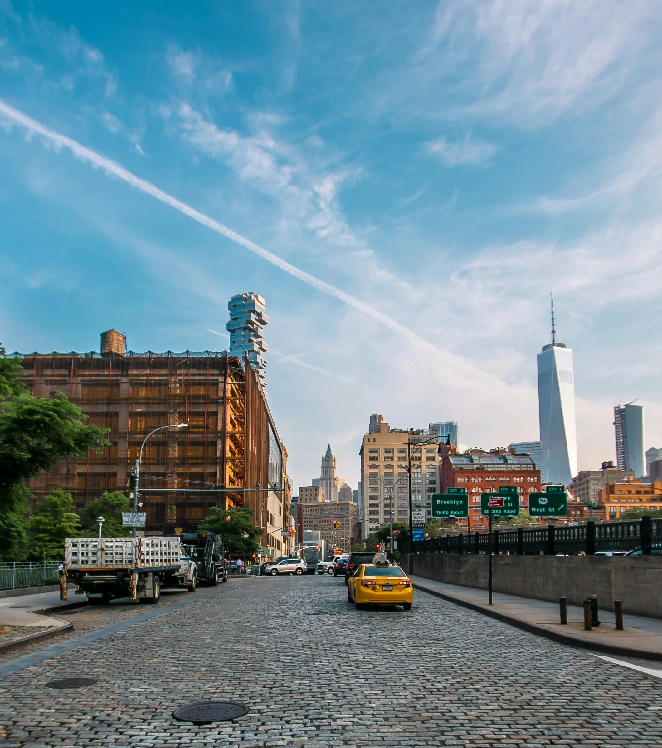 Cobblestone street in Tribeca, New York with a yellow taxi driving toward Lower Manhattan skyline, including One World Trade Center, under a blue sky with wispy clouds.