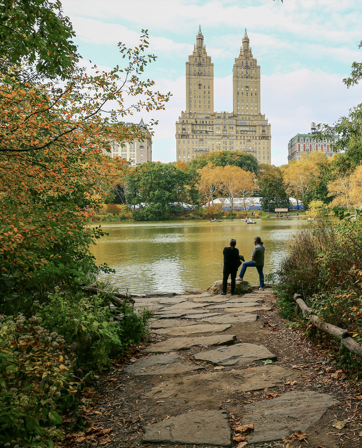 Autumn scene in Upper West Side Central Park with a rocky path leading to the water, two visitors standing at the edge of the pond, colorful fall foliage, and tall historic apartment buildings on the skyline.