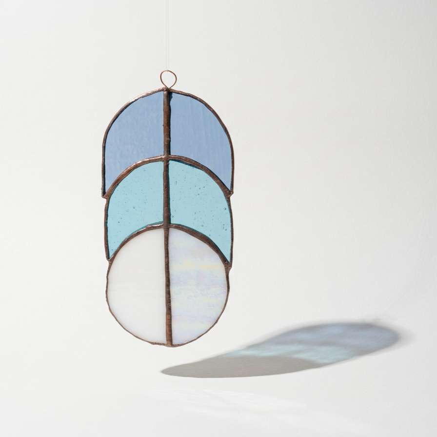 A hanging stained glass suncatcher with four semi-circular colored glass pieces in shades of blue, white, and iridescent, casting a shadow on a white background.