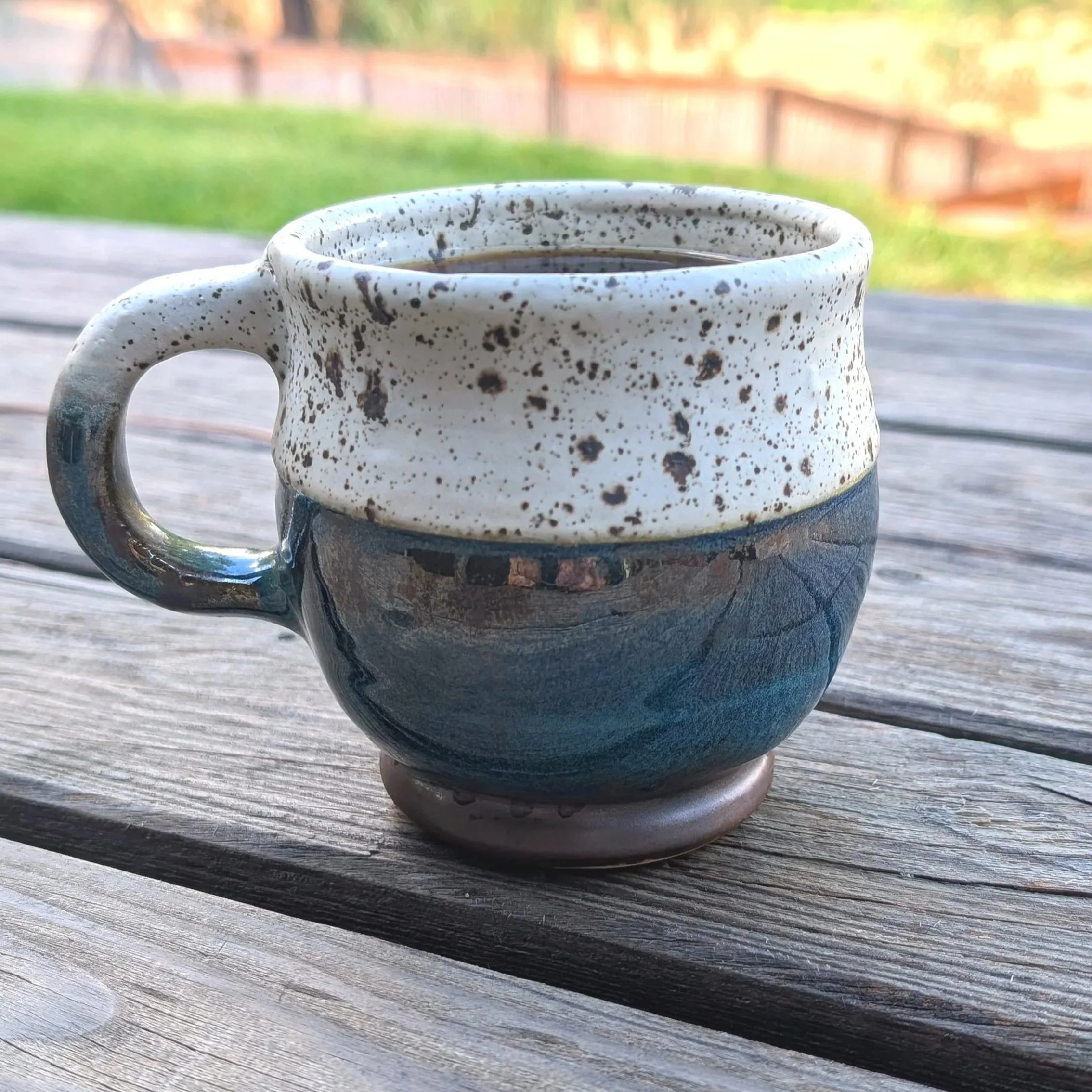 Close-up of a speckled ceramic coffee mug filled with coffee, placed on a weathered wooden surface with a blurred garden and wooden fence in the background.