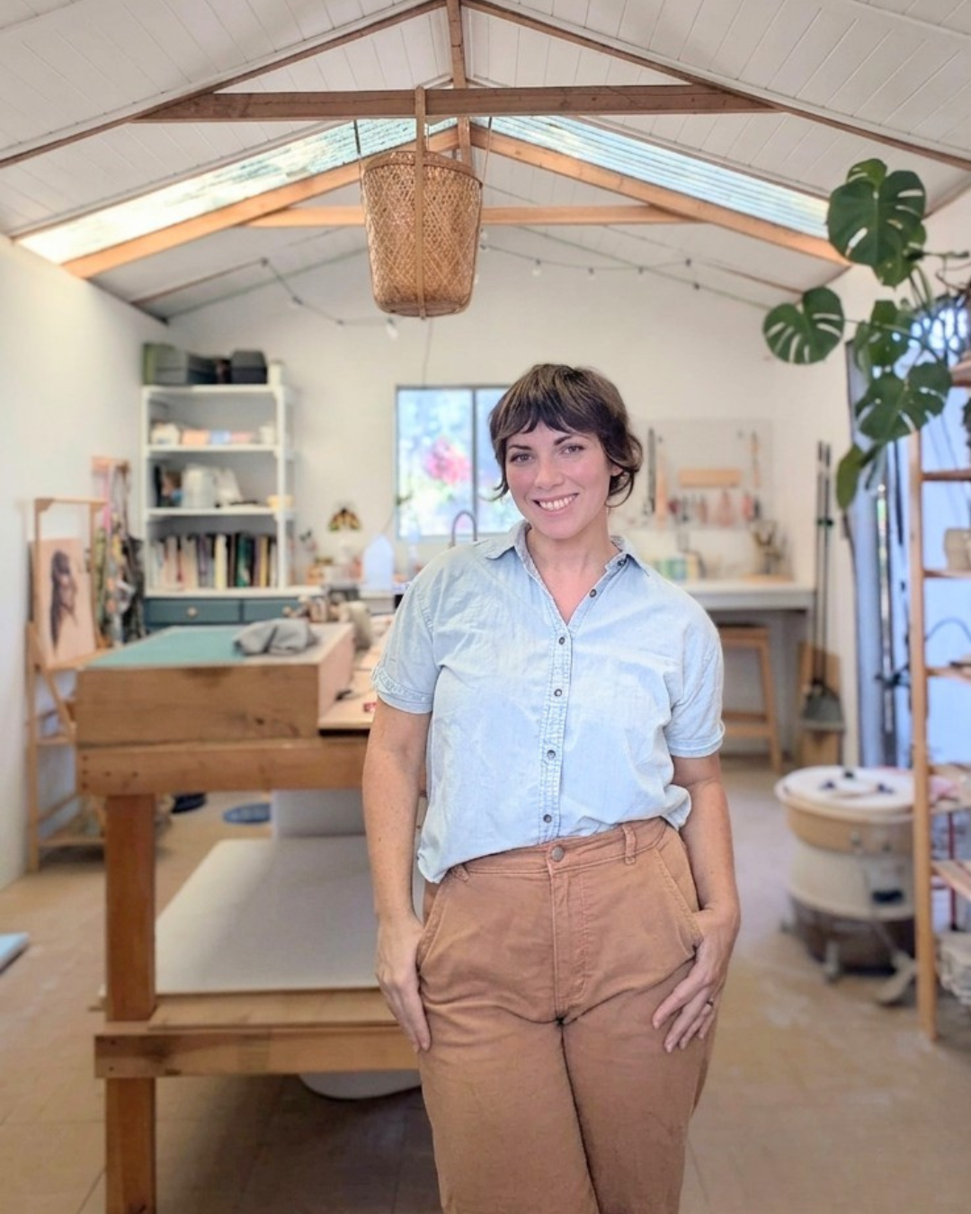 A woman standing in a well-lit, organized craft or pottery studio with shelves and worktables, smiling at the camera.