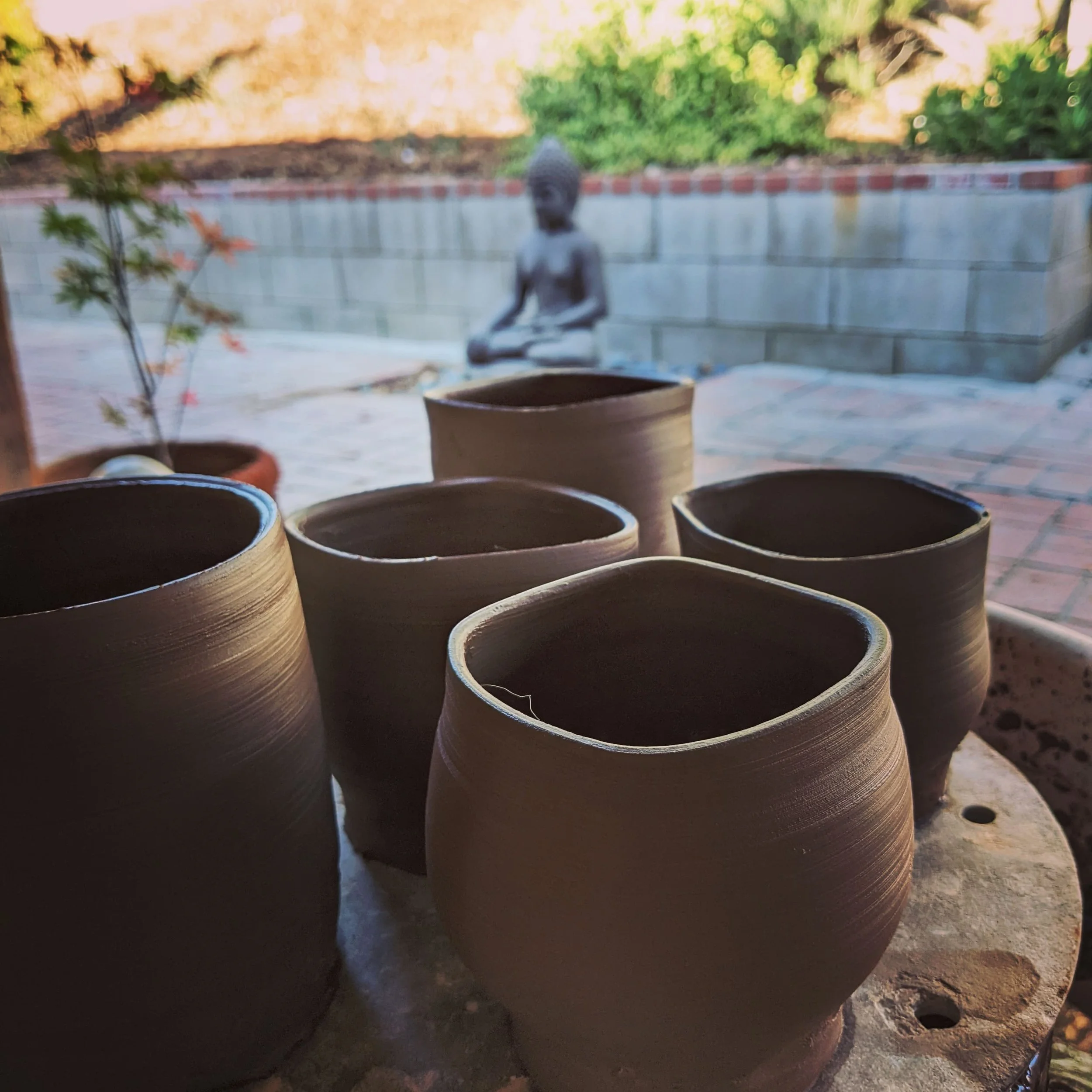 Close-up of multiple unglazed ceramic cups or pots on a rough outdoor surface, with a blurred background including greenery and a silver Buddha statue.