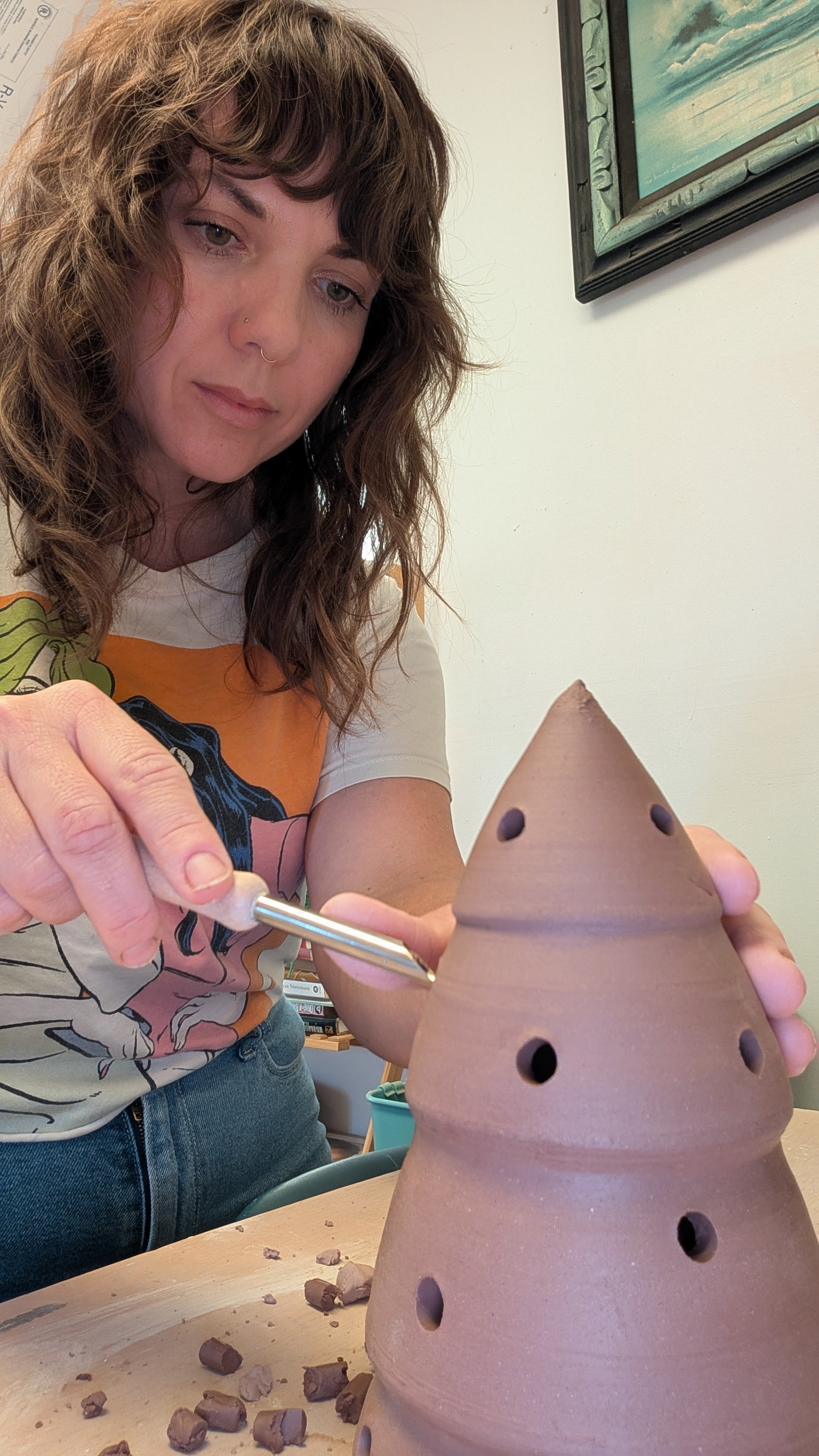 A woman with wavy brown hair and a nose piercing is decorating a tall, cone-shaped, unglazed terracotta pottery piece with multiple holes. She is holding a tool and working on the pottery, which has a rough surface and is placed on a wooden table, with small clay fragments scattered around.