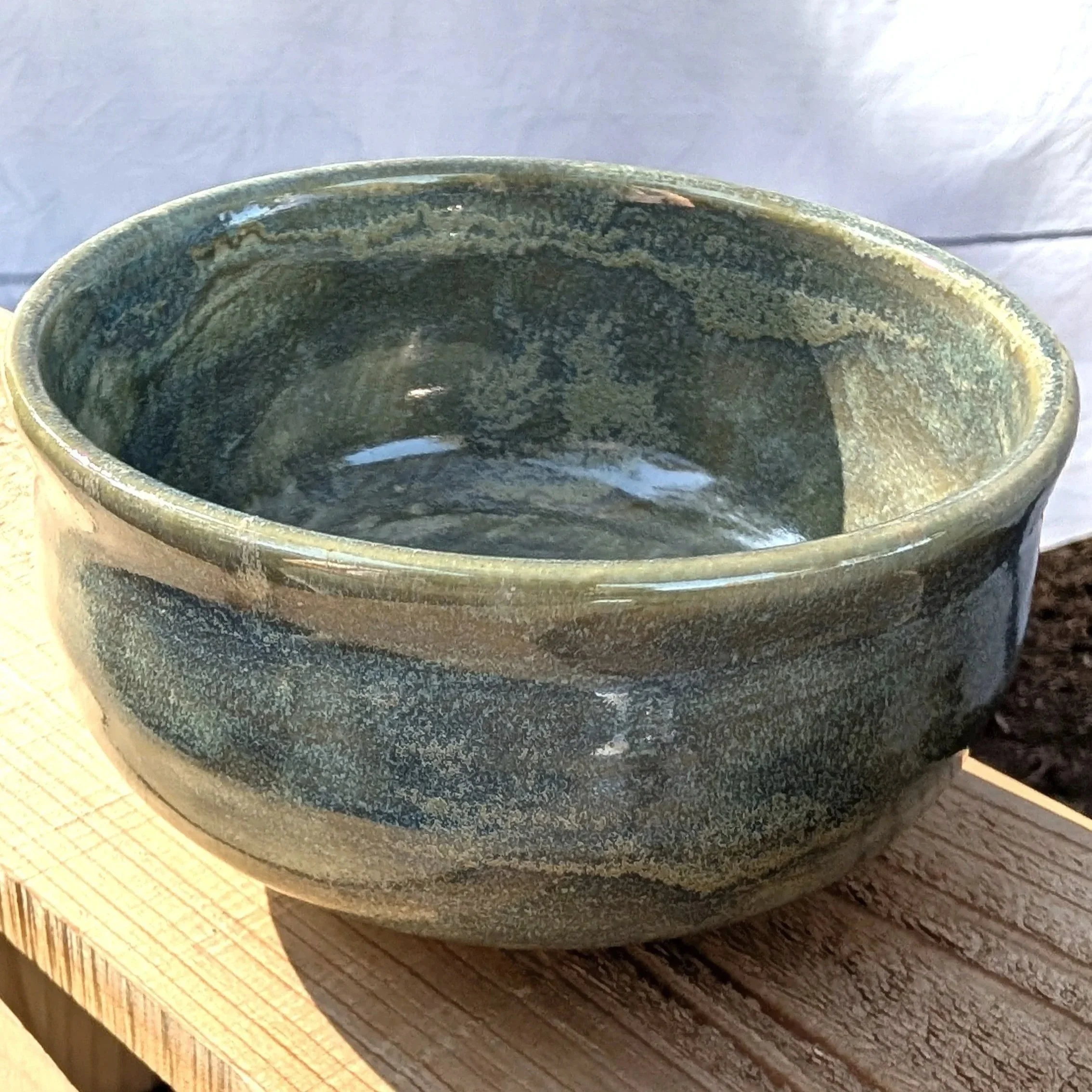 A close-up of a glazed ceramic pot with a greenish-blue glaze, sitting on a wooden surface outdoors.