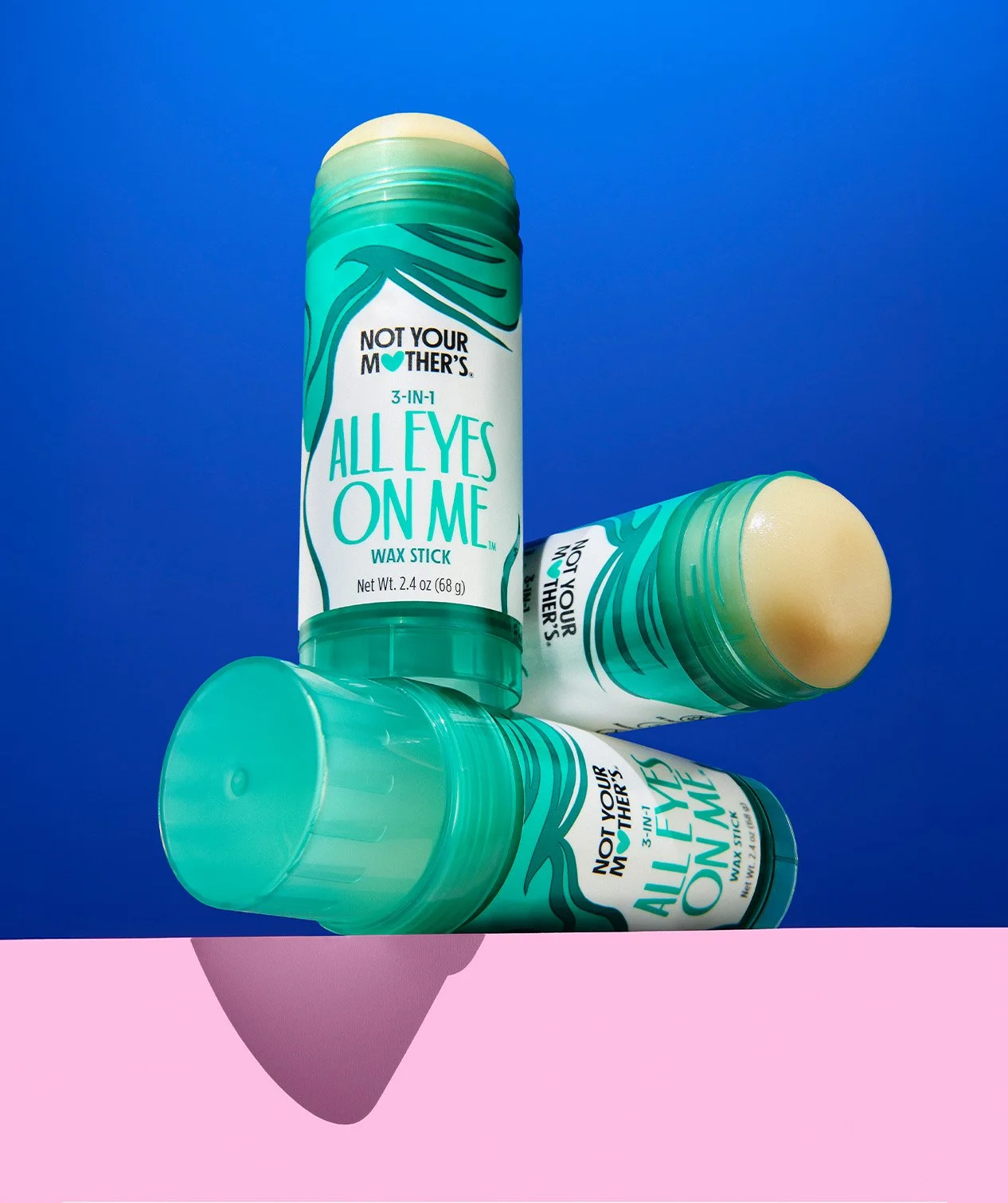 Three Not Your Mother's All Eyes On Me Wax sticks with colorful packaging, one standing upright, two lying horizontally, against a blue background with pink surface, casting a shadow.