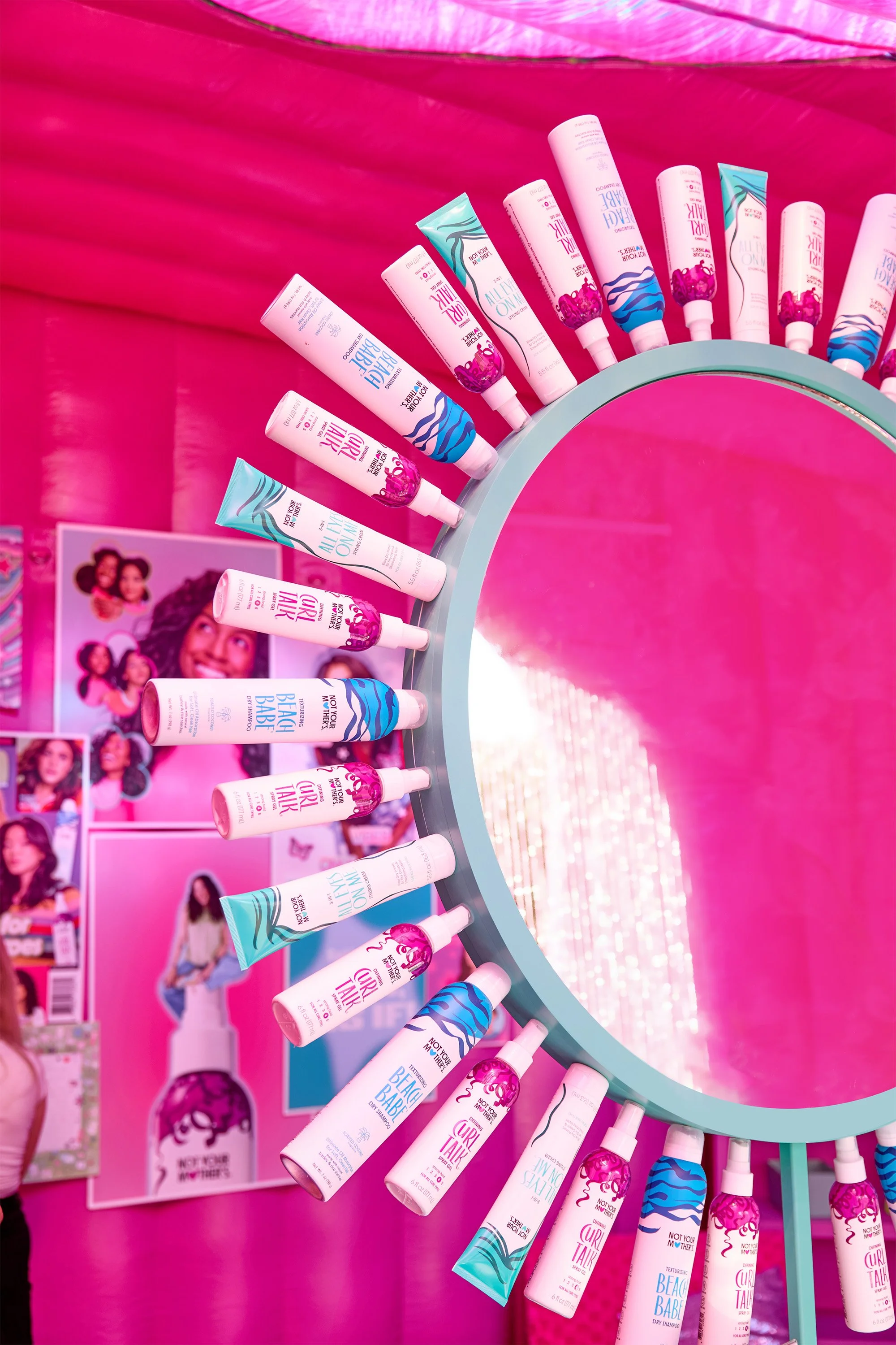 A round mirror with hair care product bottles arranged around its edge, set against a vibrant pink background with promotional posters attached behind.