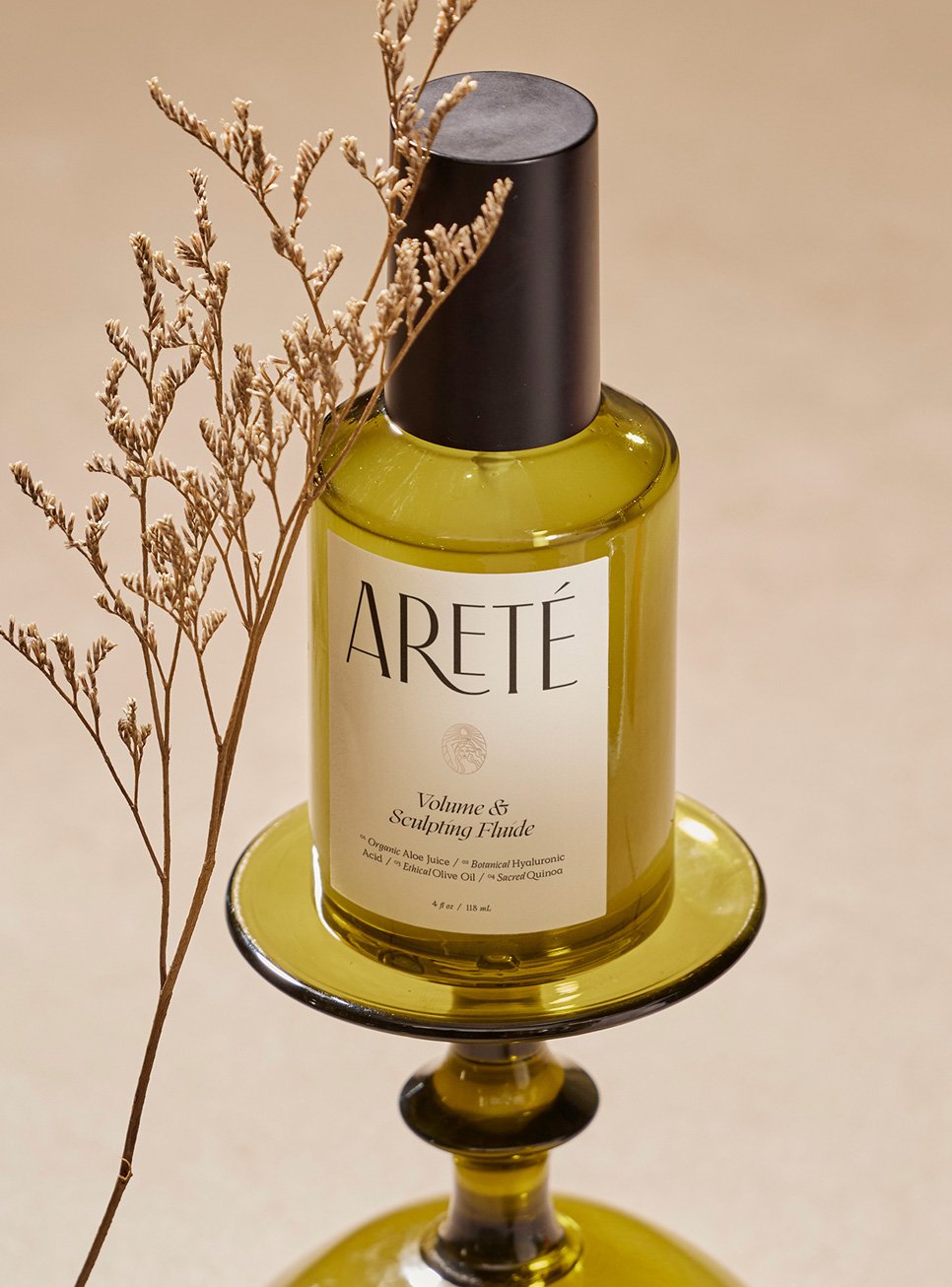 A bottle labeled 'ARETÉ Volume & Sculpting Fluid' with ingredients including organic aloe juice, botanical hyaluronic acid, olive oil, and sacred quinia, placed on a decorative yellow glass stand with a dried branch nearby.