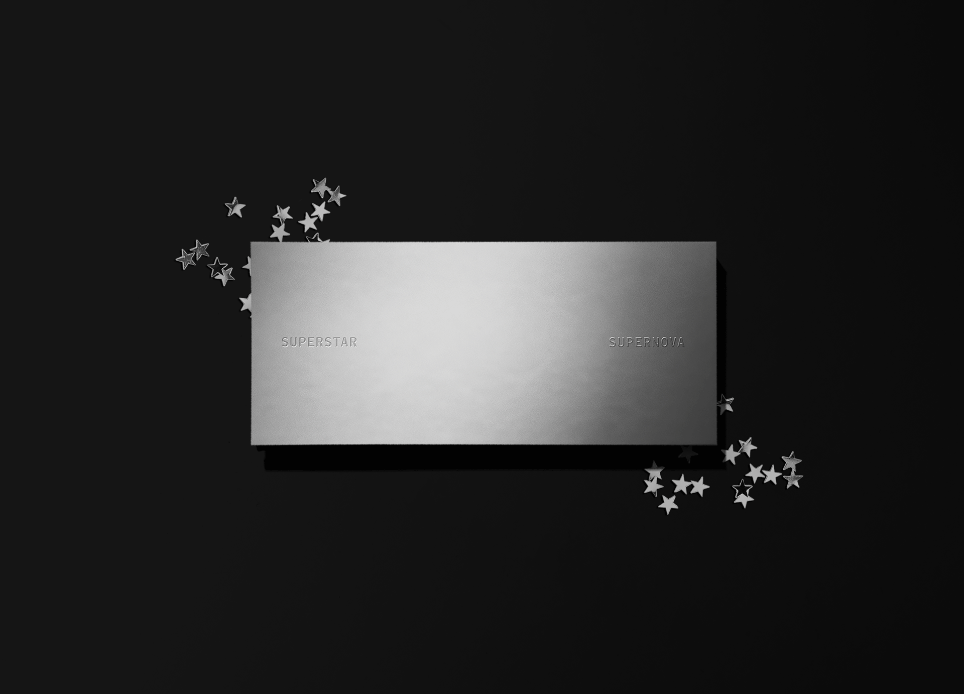 A silver rectangular box with words 'SUPERSTAR' and 'SUPERNOVA' engraved on it, surrounded by small silver star-shaped confetti on a black background.