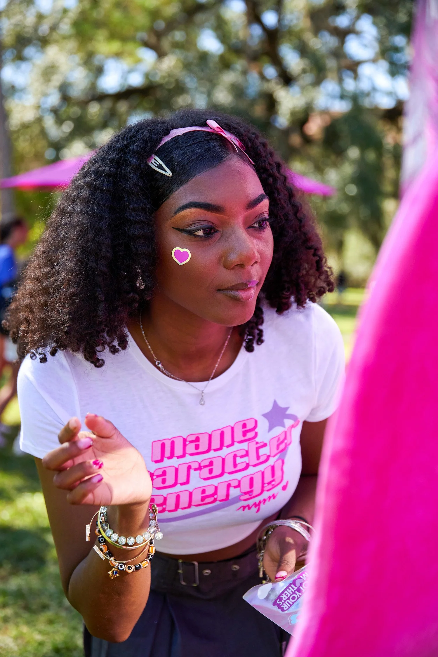 A woman with curly hair, wearing a white t-shirt with pink text, accessorized with jewelry, and adorned with a heart-shaped sticker on her cheek, is outdoors in a park setting.