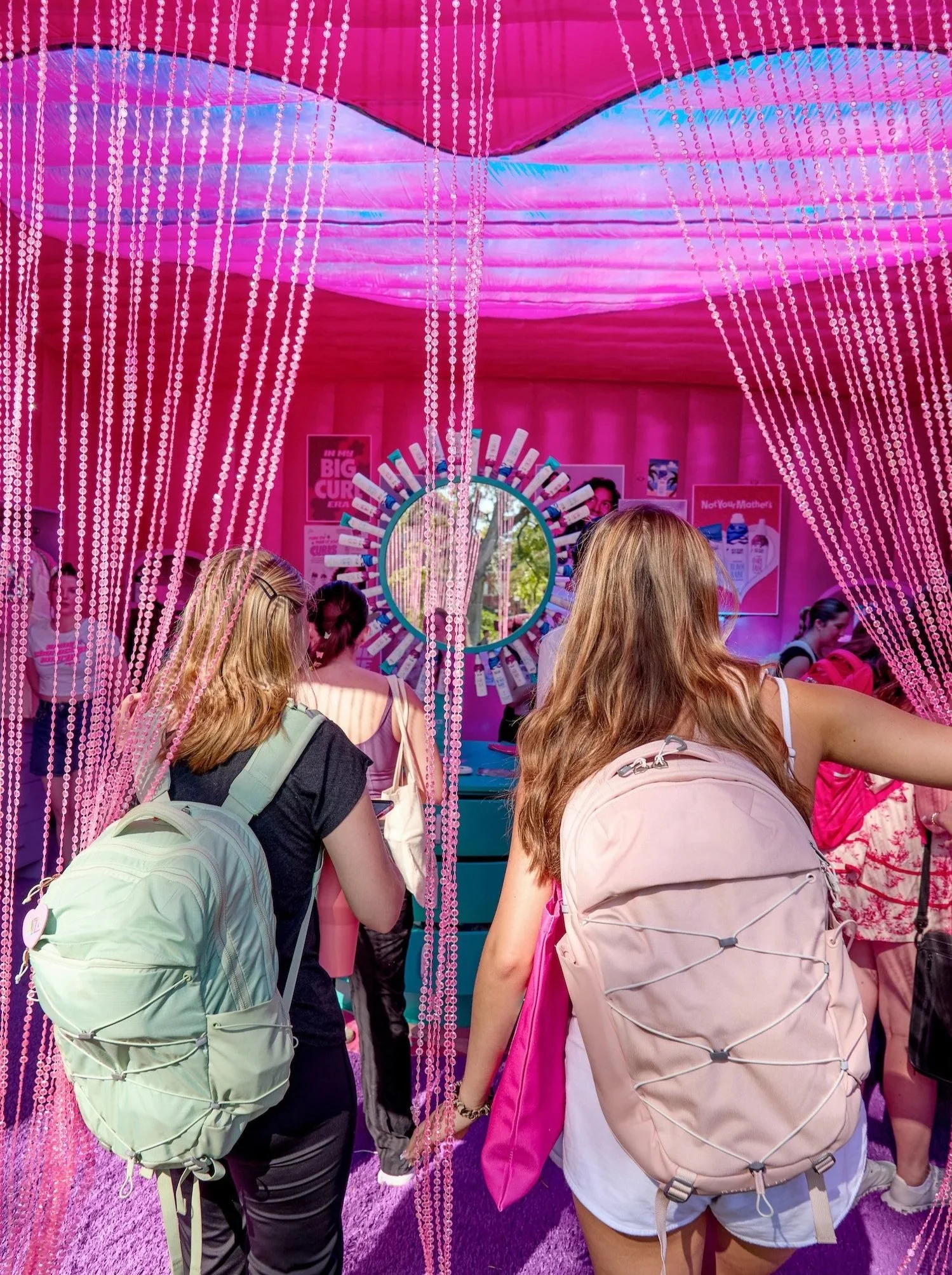 People at a pink-themed event or exhibit with hanging beads, a circular display, and posters on the walls.