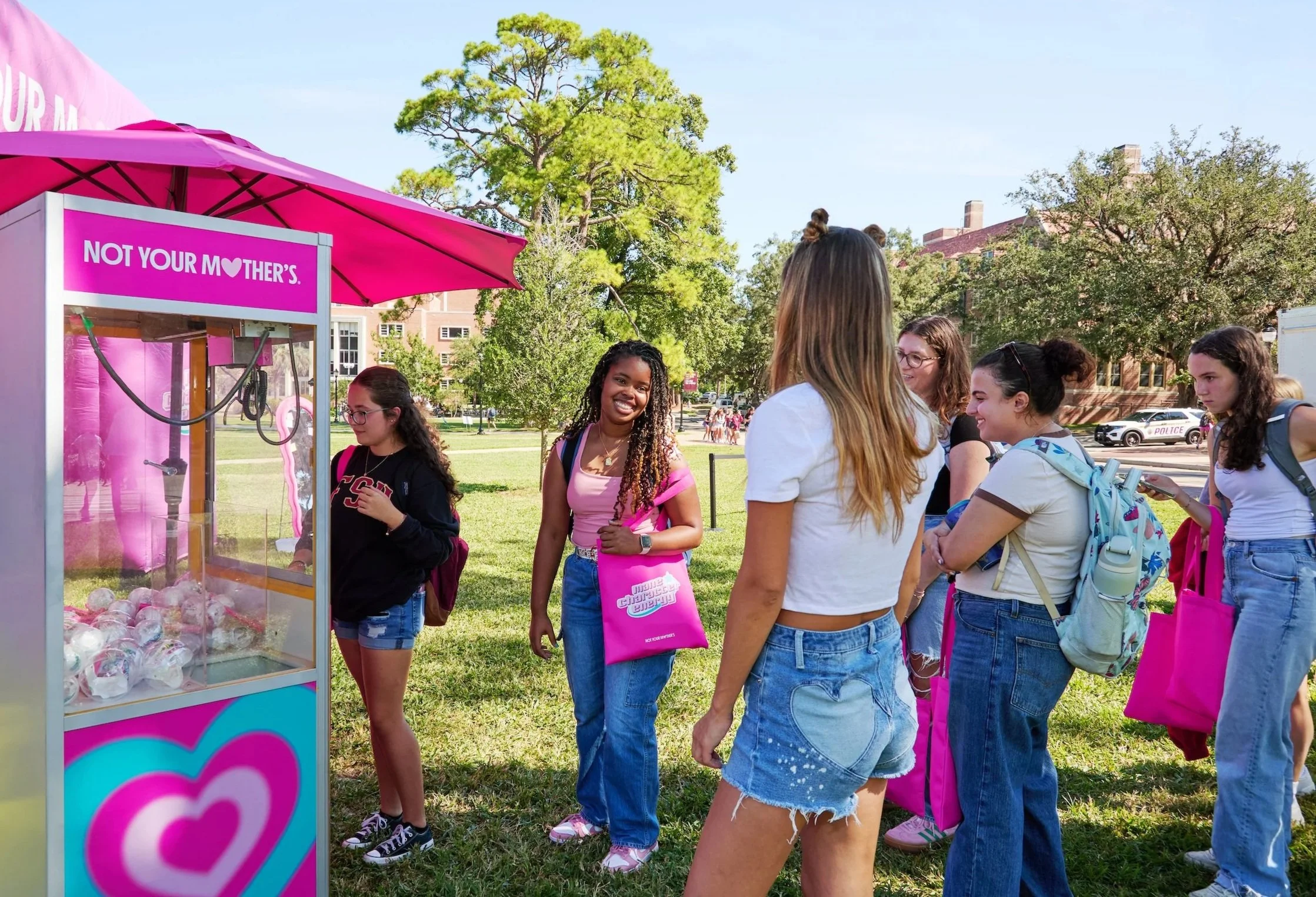 Group of young girls standing in line at a pink claw machine labeled 'Not Your Mother’s' on a sunny day, with trees and a building in the background.