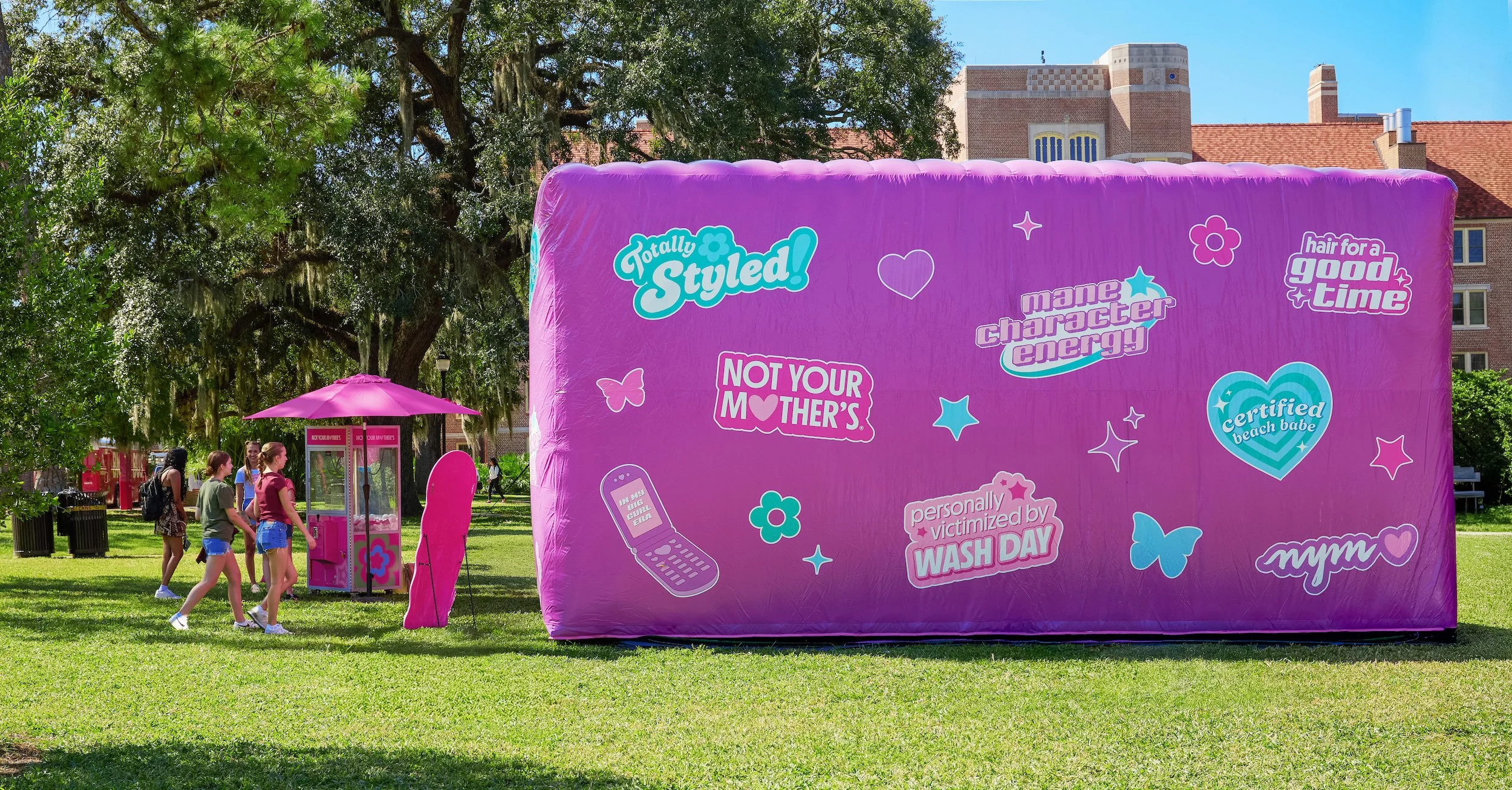 A pink inflatable structure with various stickers and phrases, set up outdoors in a park-like area with trees and buildings. Next to it, four girls are walking past pink vending machines with an umbrella providing shade.