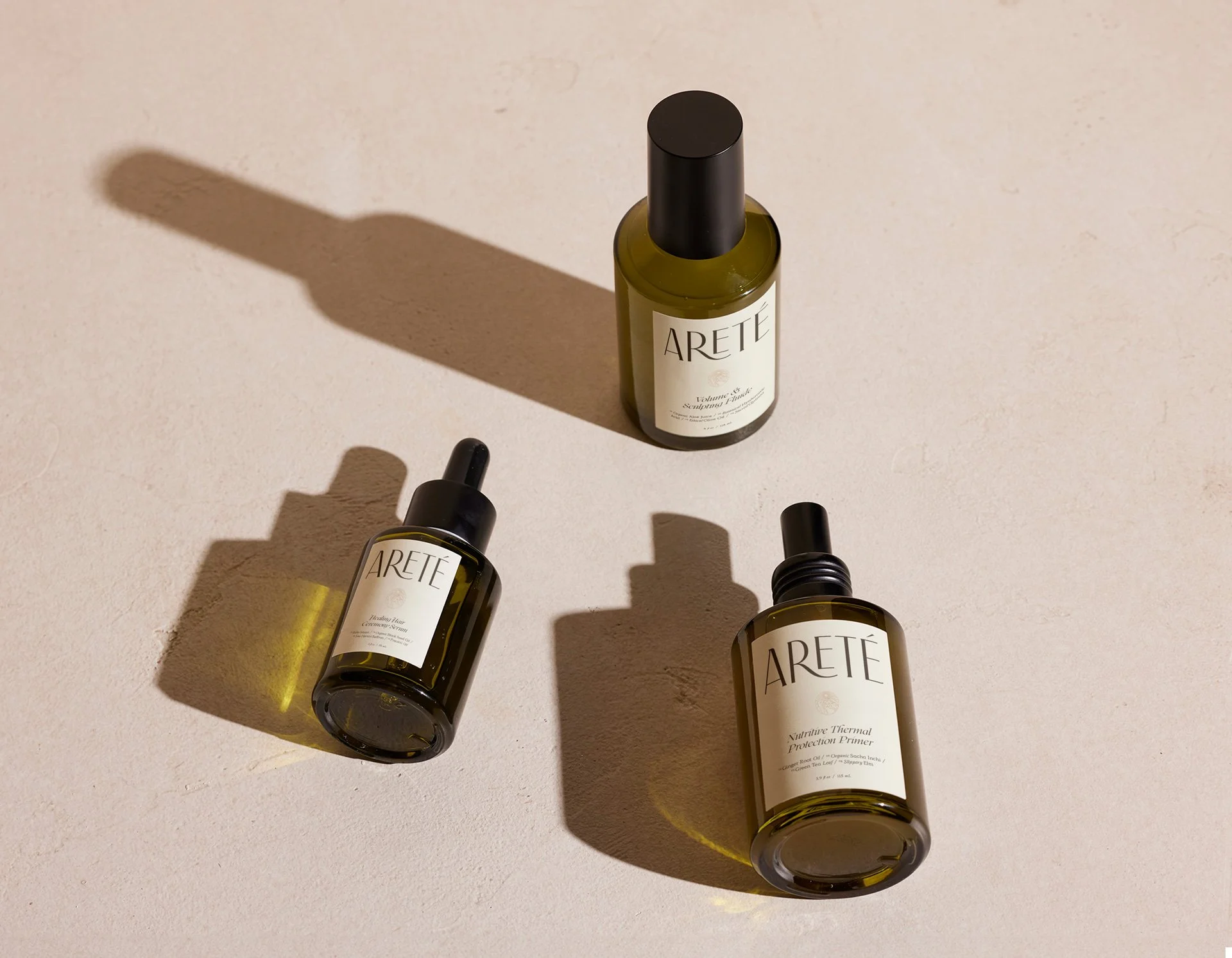 Three bottles of ARETÉ skincare products on a beige surface, casting shadows in different directions.