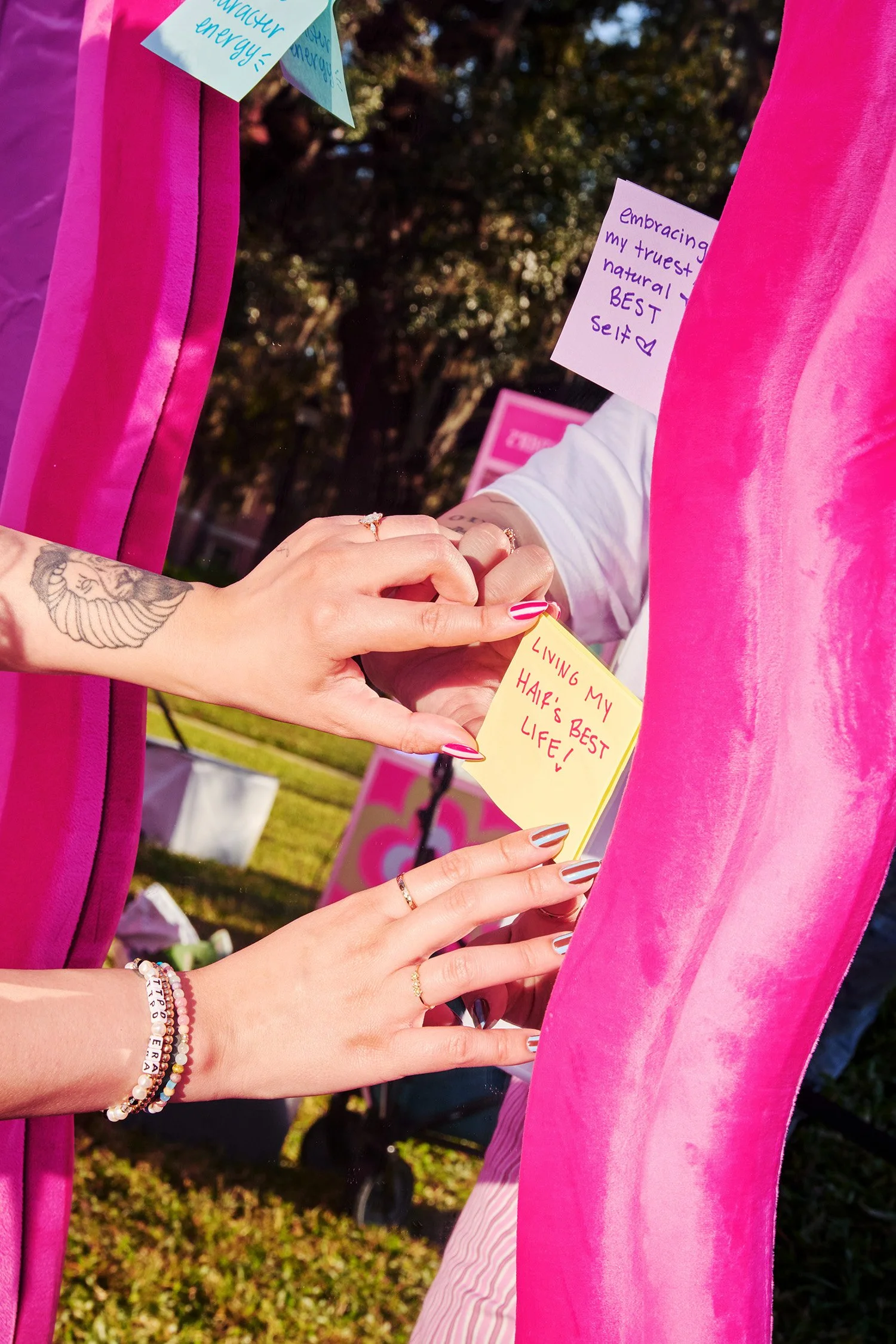 People writing handwritten notes on sticky notes and attaching them to a pink velvet-covered mirror at an outdoor event. One note reads 'LIVING MY HAIR'S BEST LIFE!' and other notes contain positive affirmations.